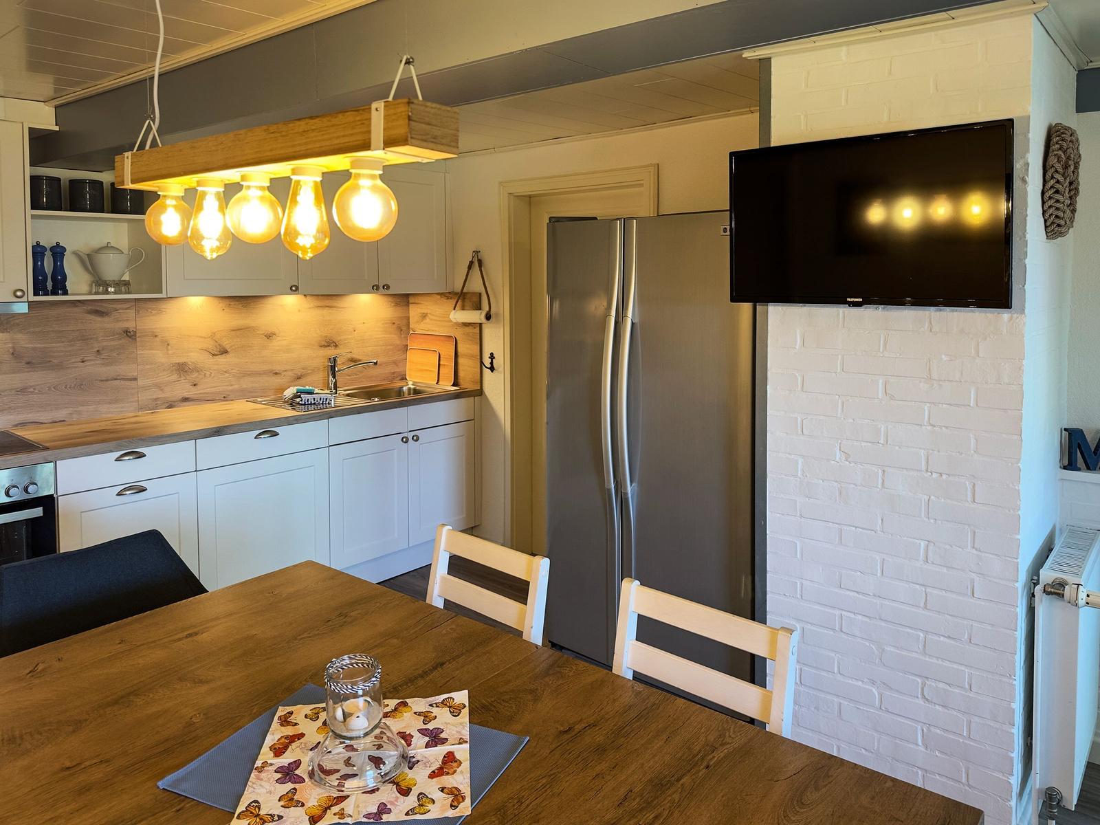 Küche mit Essbereich, Kühlschrank, TV und Holztisch mit Stühlen.