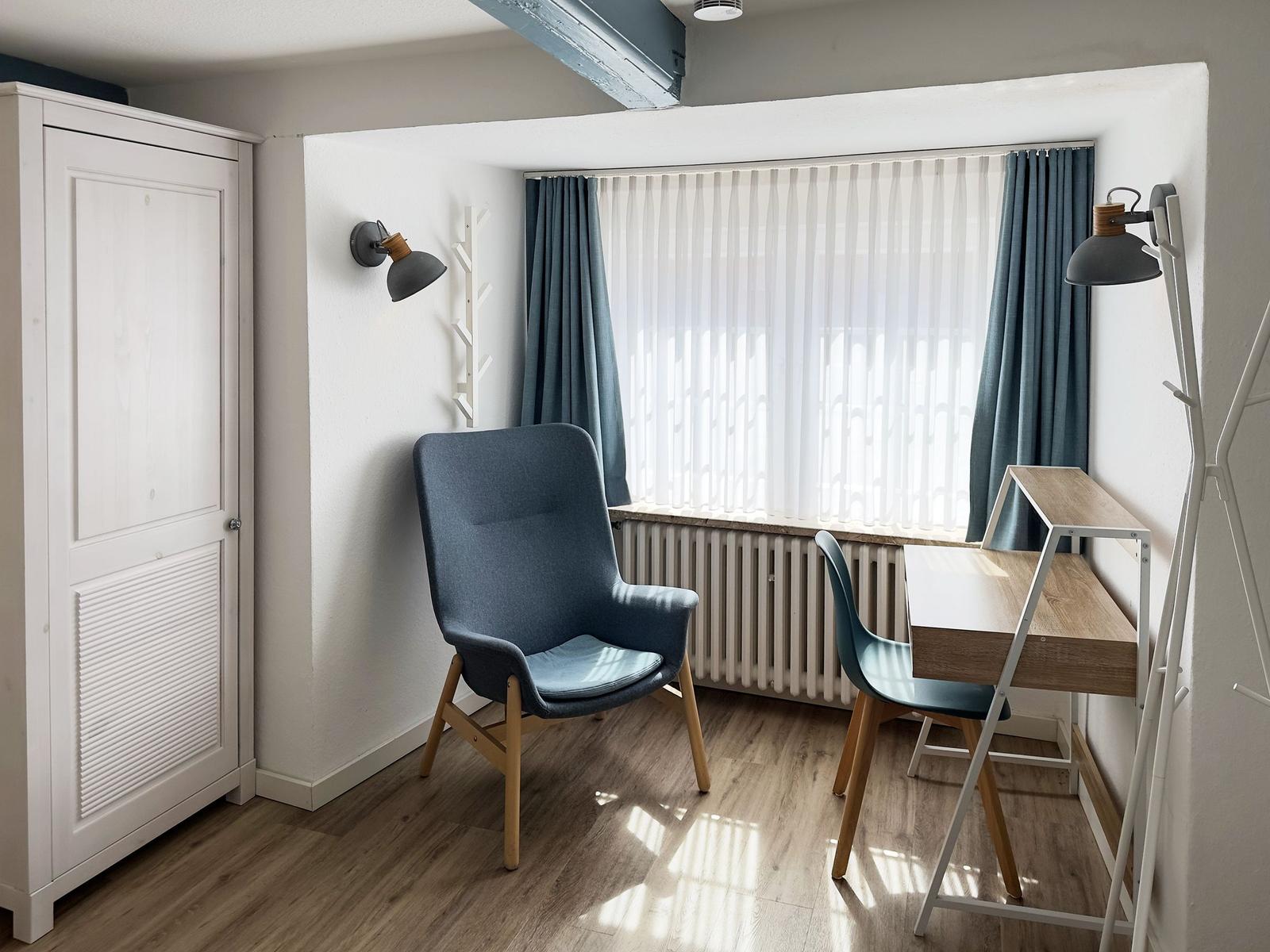 Ein Raum mit Schreibtisch, Stuhl und Kleiderschrank neben großem Fenster.
