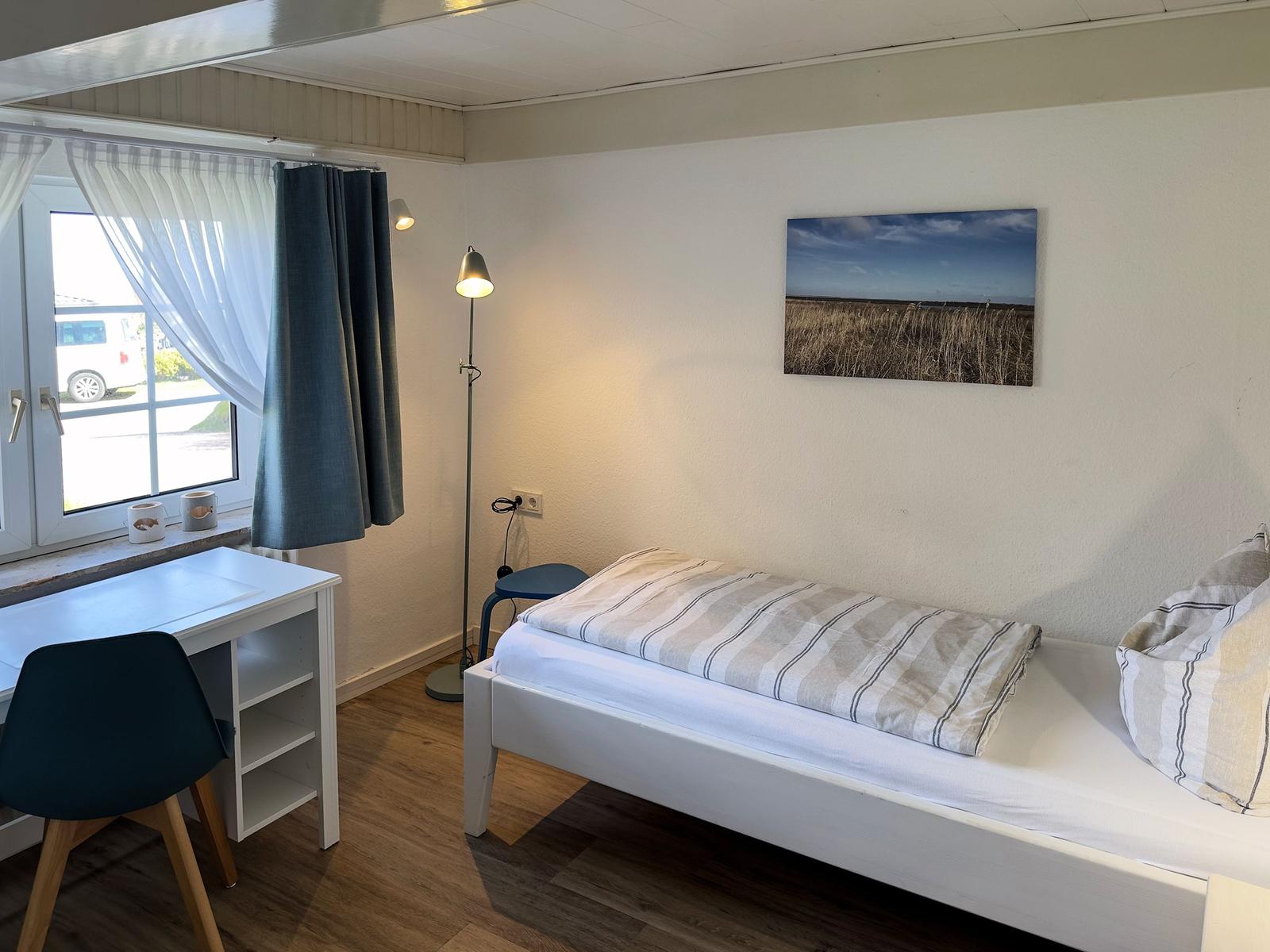 Ein Schlafzimmer mit Bett, Schreibtisch und Fenster. Wandbild und Stehlampe.