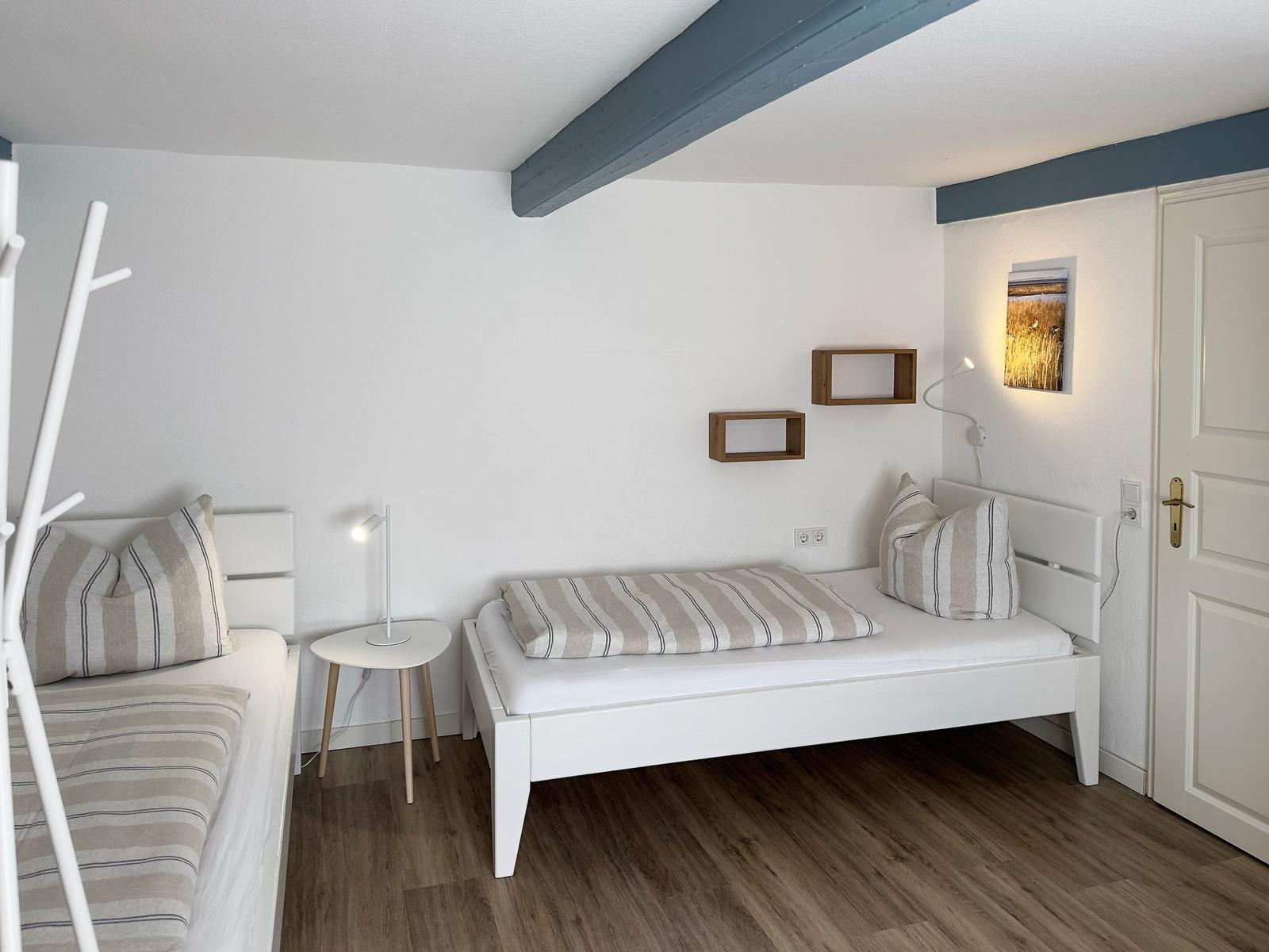 Zimmer mit zwei Betten, Holzboden und Wandregalen. Deckenbalken und Wandbild sichtbar.