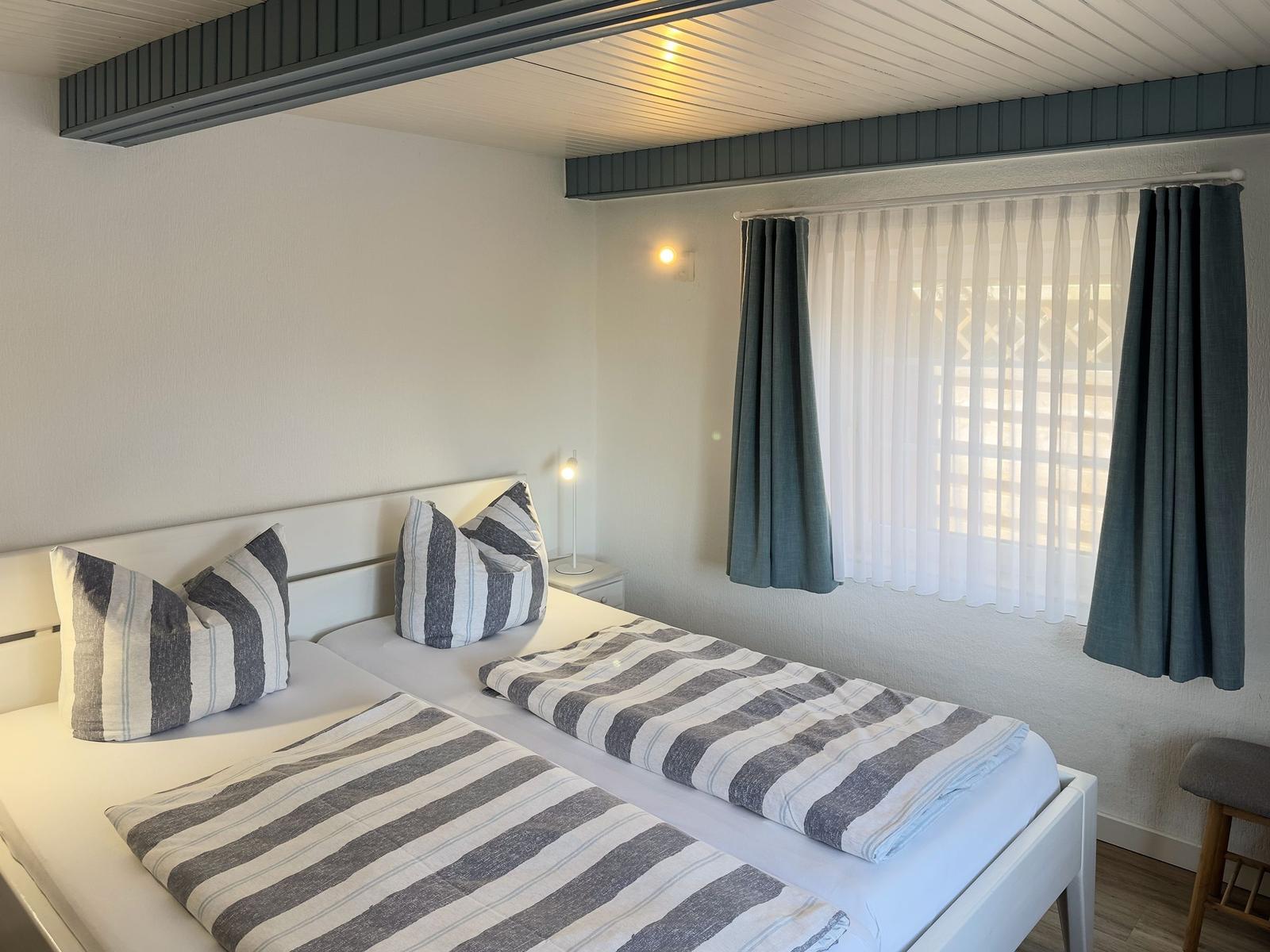 Zwei Betten mit gestreiften Bettwäsche, Fenster mit Vorhängen und Nachttisch mit Lampe.