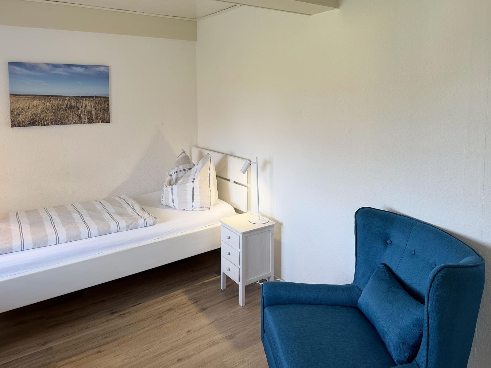 Ein Schlafzimmer mit Bett, Nachttisch und blauem Sessel. Wandbild zeigt eine Landschaft.