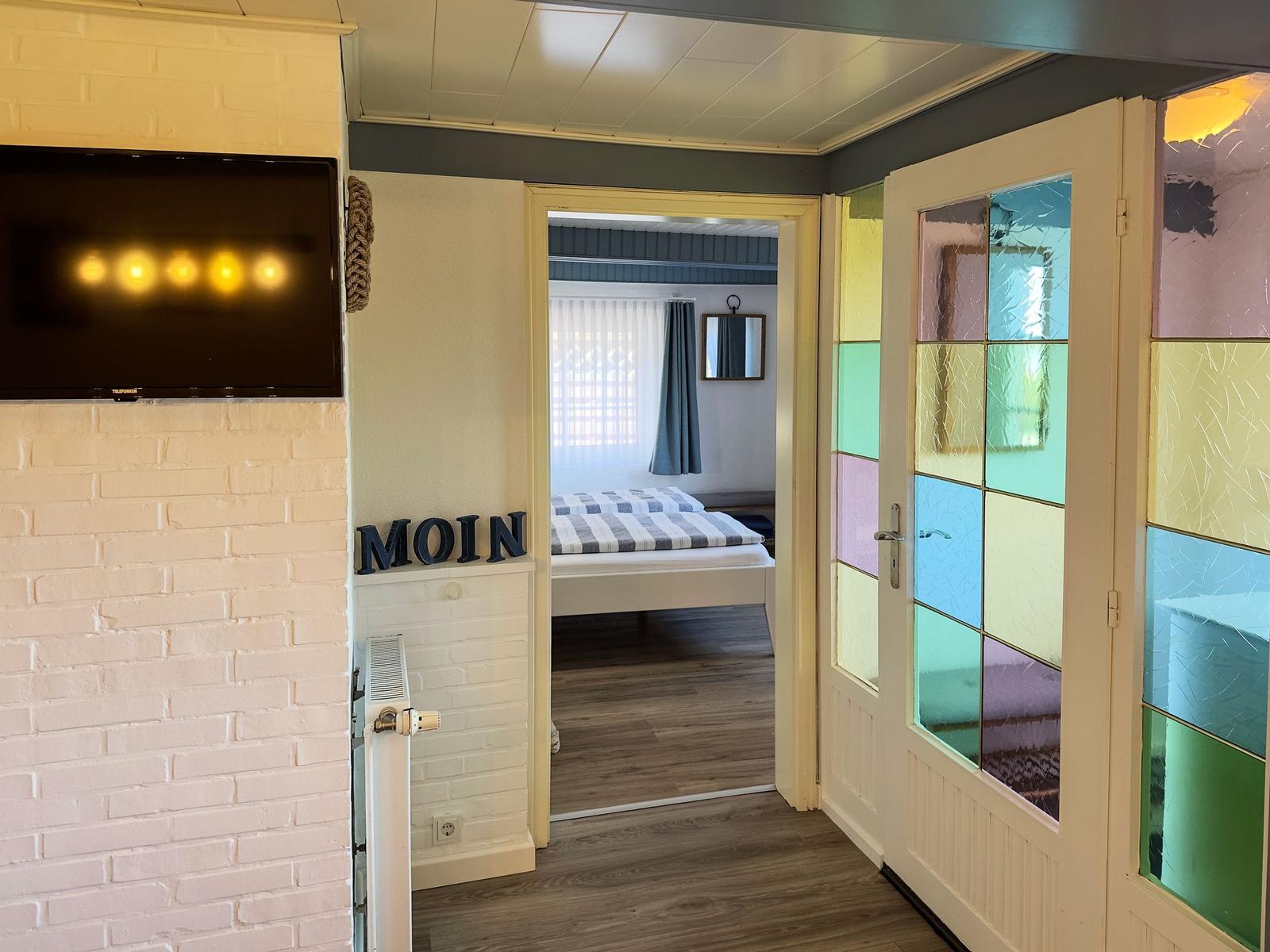 Schlafzimmer mit Bett und Fenster. Flur mit TV und bunten Glasscheiben.