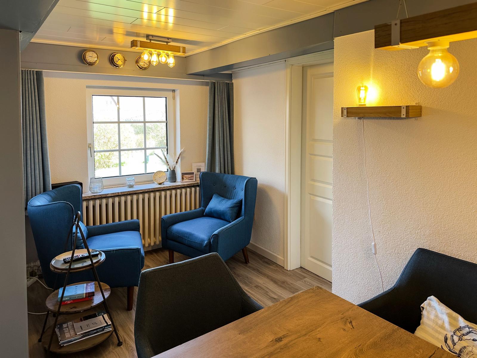 Zwei blaue Sessel stehen vor einem Fenster mit Blick nach draußen. Holztisch und Wandlampe sind sichtbar.