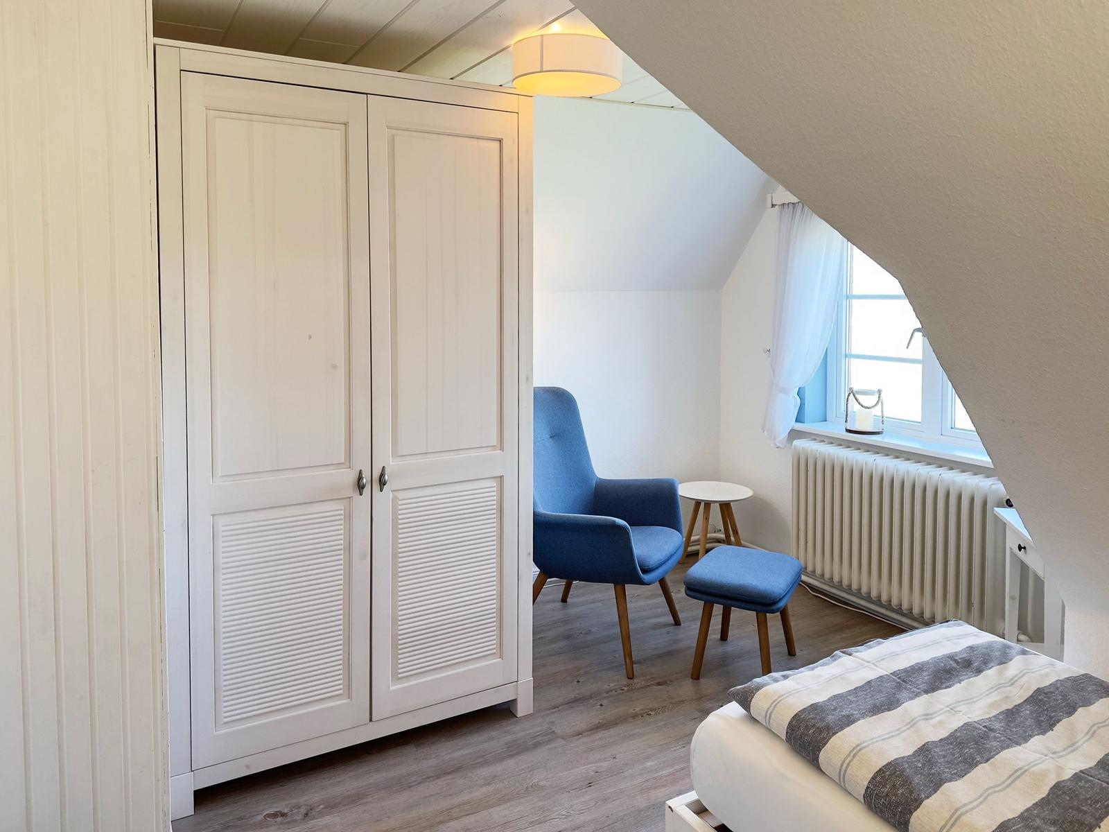 Schlafzimmer mit weißem Schrank, blauer Sitzgelegenheit und Fenster mit Vorhang.