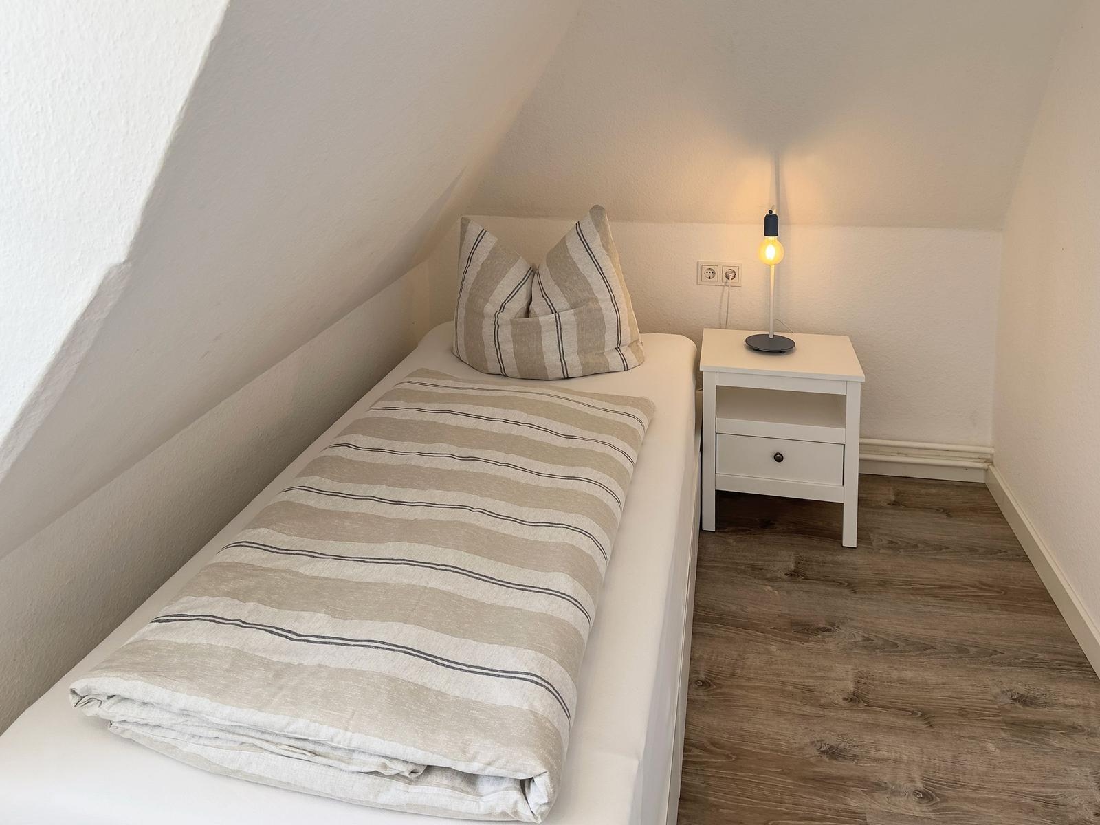 Schlafzimmer mit Einzelbett, Nachttisch und Stehlampe unter schrägen Dachdecken.