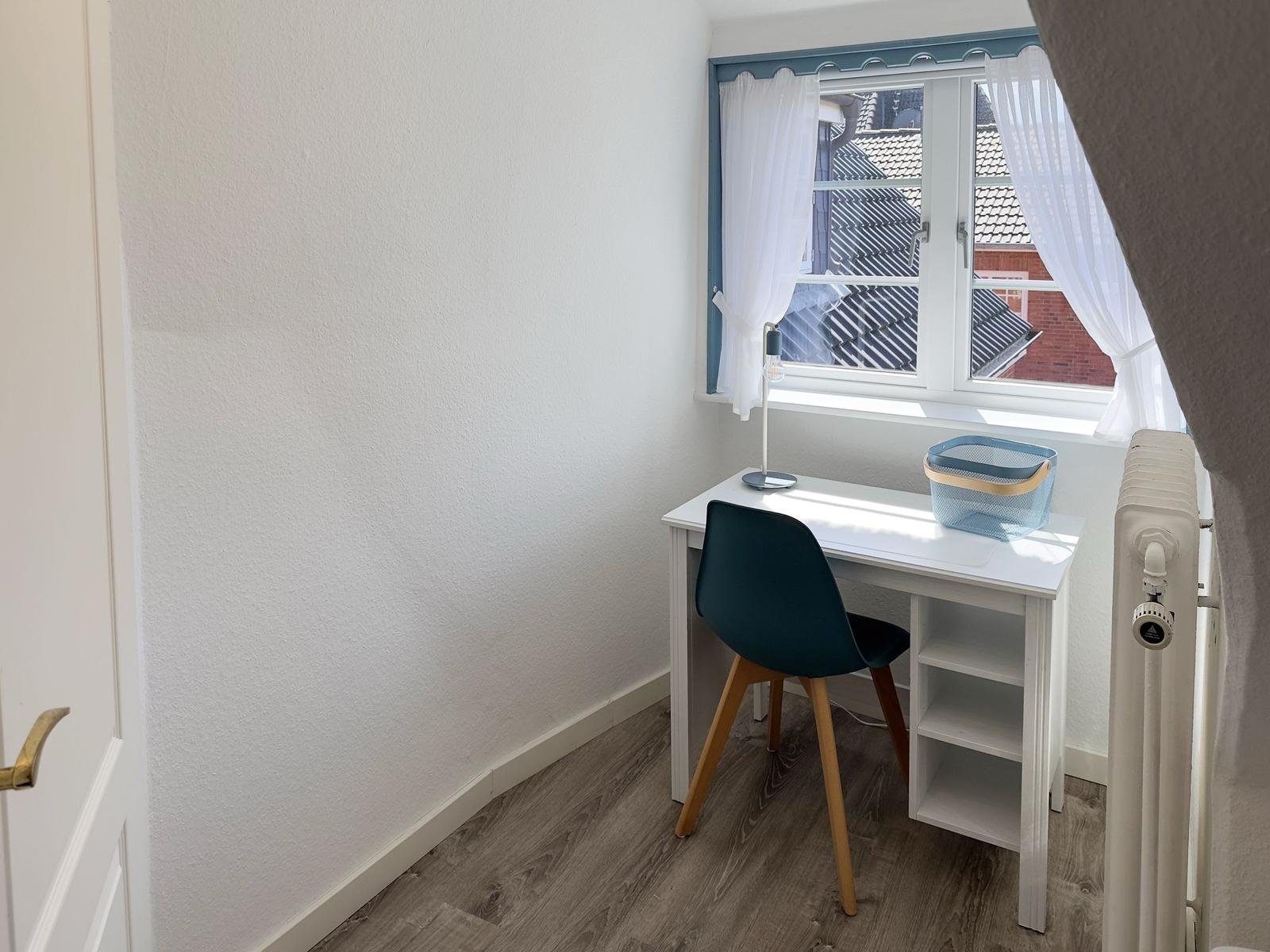 Ein Arbeitszimmer mit Schreibtisch, Stuhl und Fenster mit weißen Vorhängen.