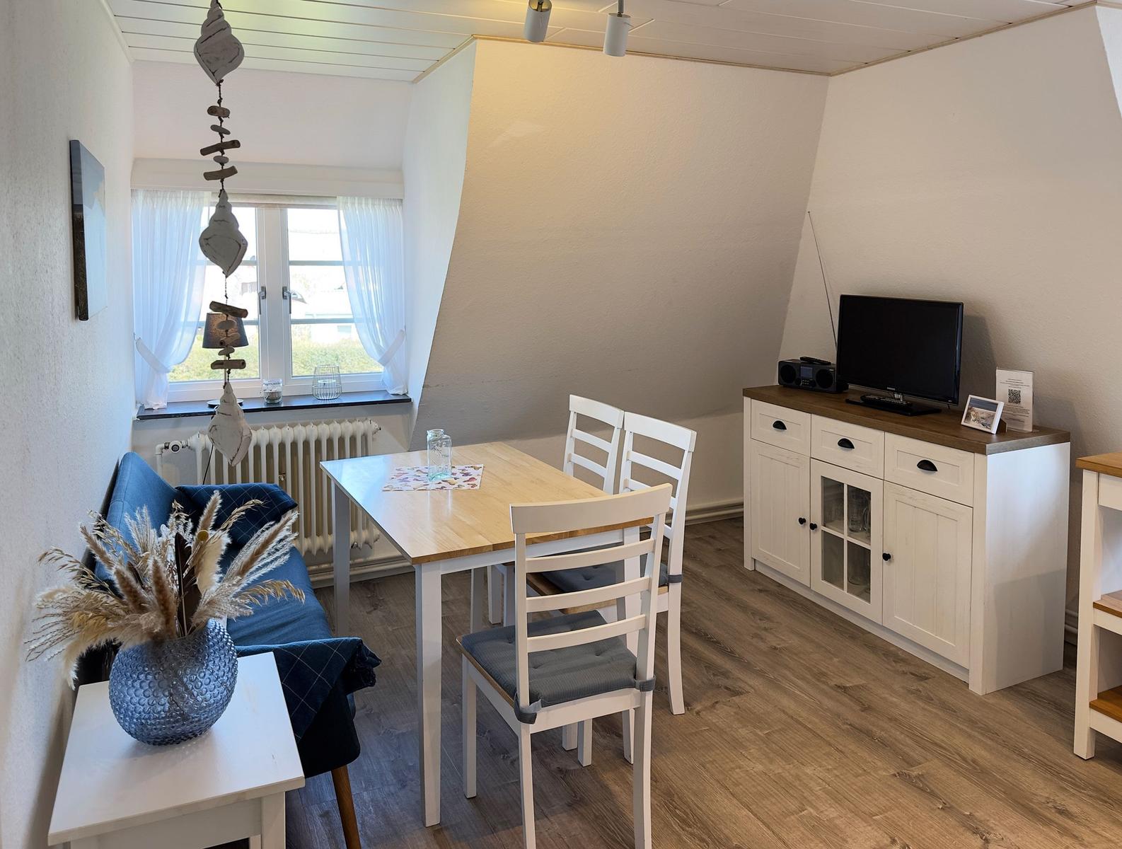 Essbereich mit Tisch, Stühlen, Sofa und Fernseher. Holzboden und Fenster mit Vorhängen.