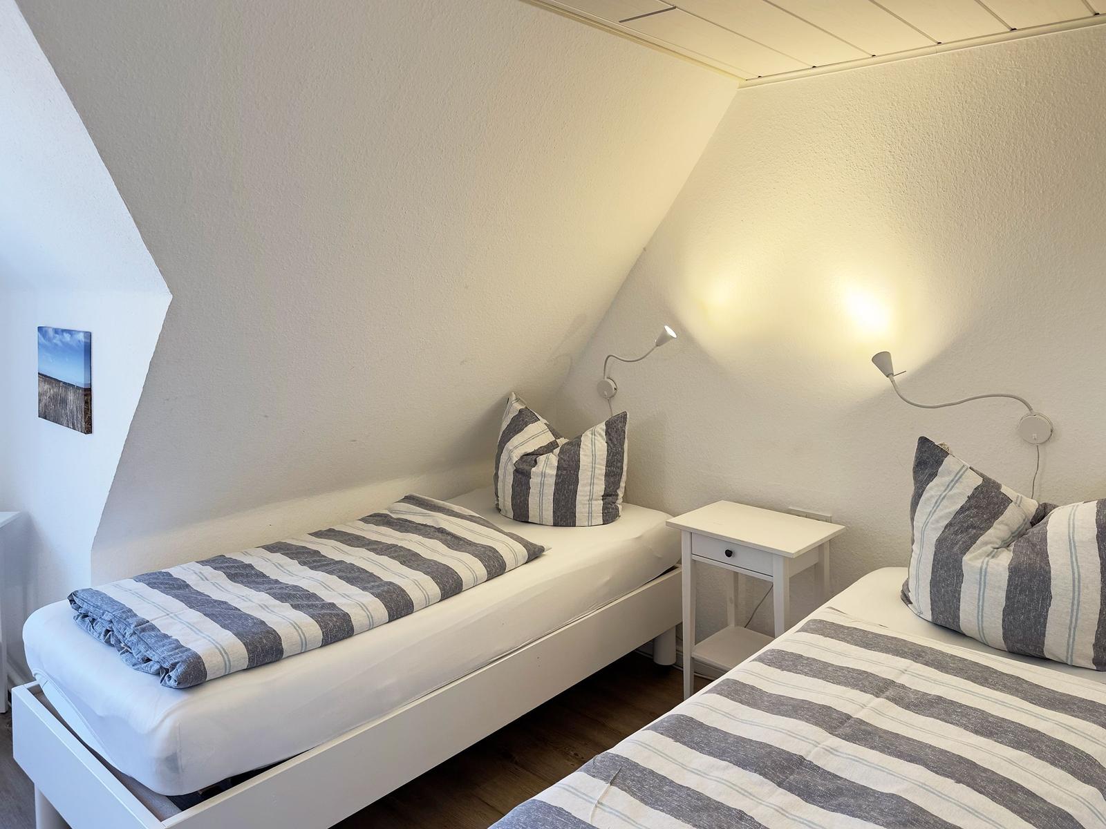 Zwei Einzelbetten mit Streifenbezug, Nachttisch und Wandlampen in einem Dachzimmer.