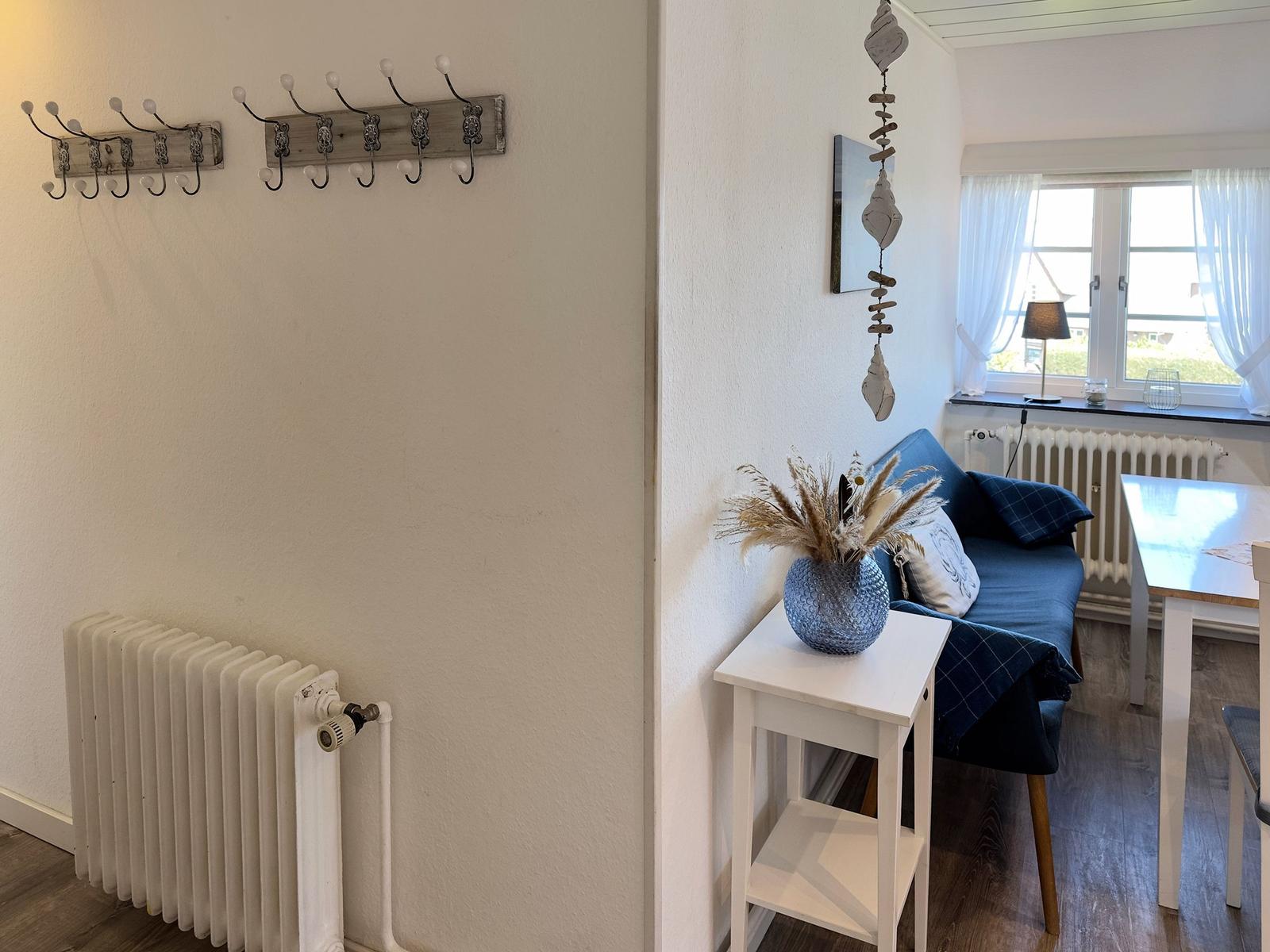 Wohnbereich mit Sofa, Tisch und Fenster. An der Wand hängen Garderobenhaken und ein Hänger.