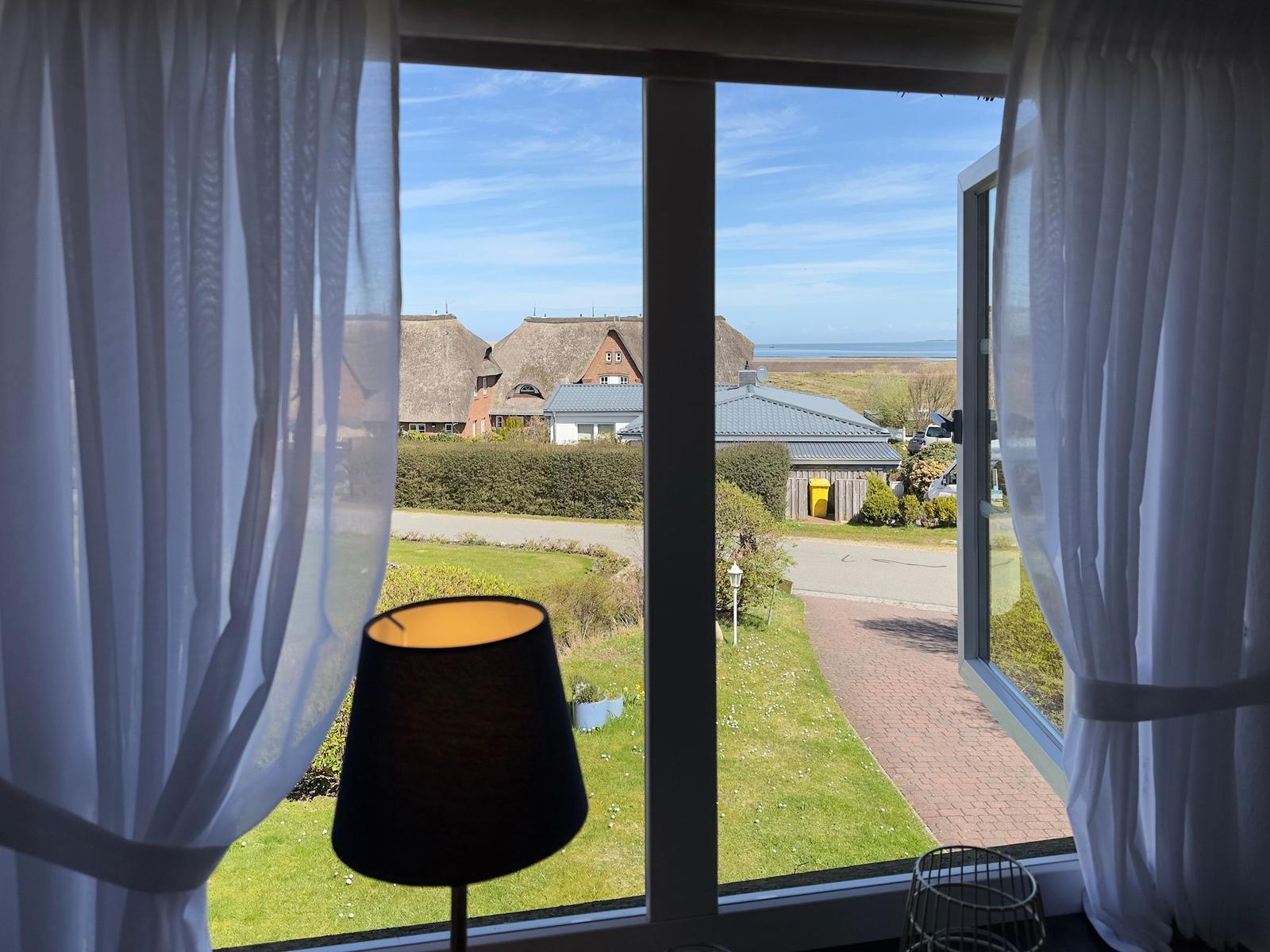 Durch das Fenster blickt man auf eine grüne Wiese, Häuser und das Meer.