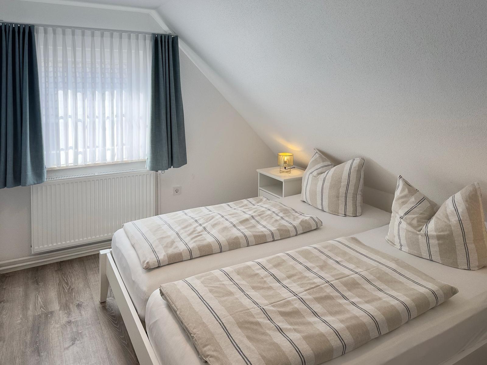 Room with two beds, window with curtains, and bedside table with lamp.