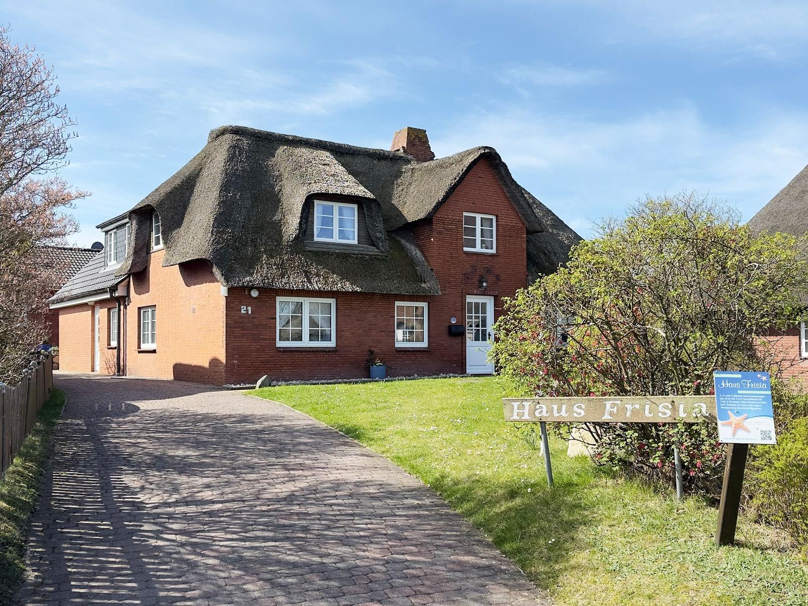 Red house with thatched roof and white windows. A sign with the name Haus Frisia stands in front of the house.