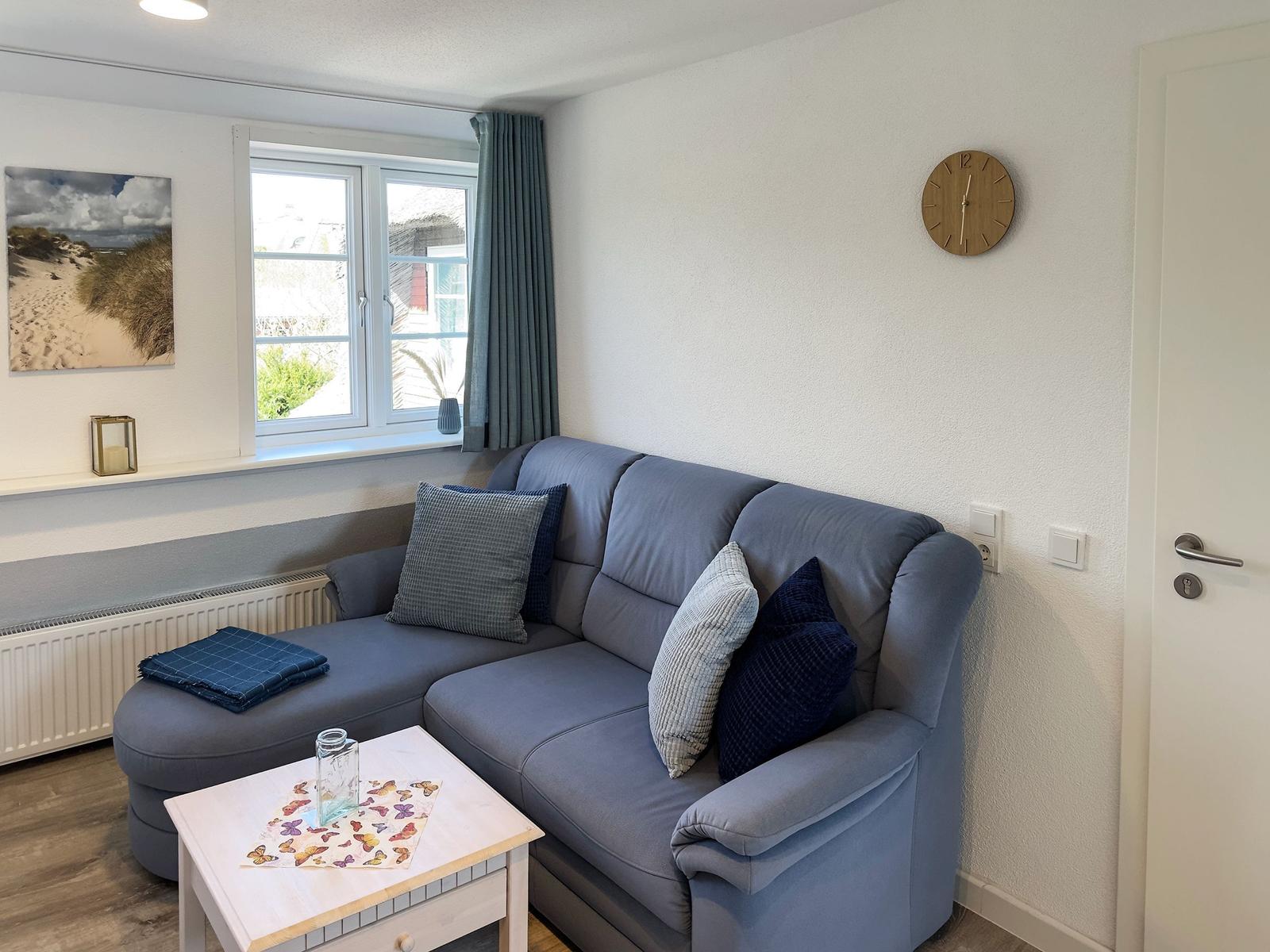 Living room with gray sofa, table, and wall clock. Window with curtains and wall picture.