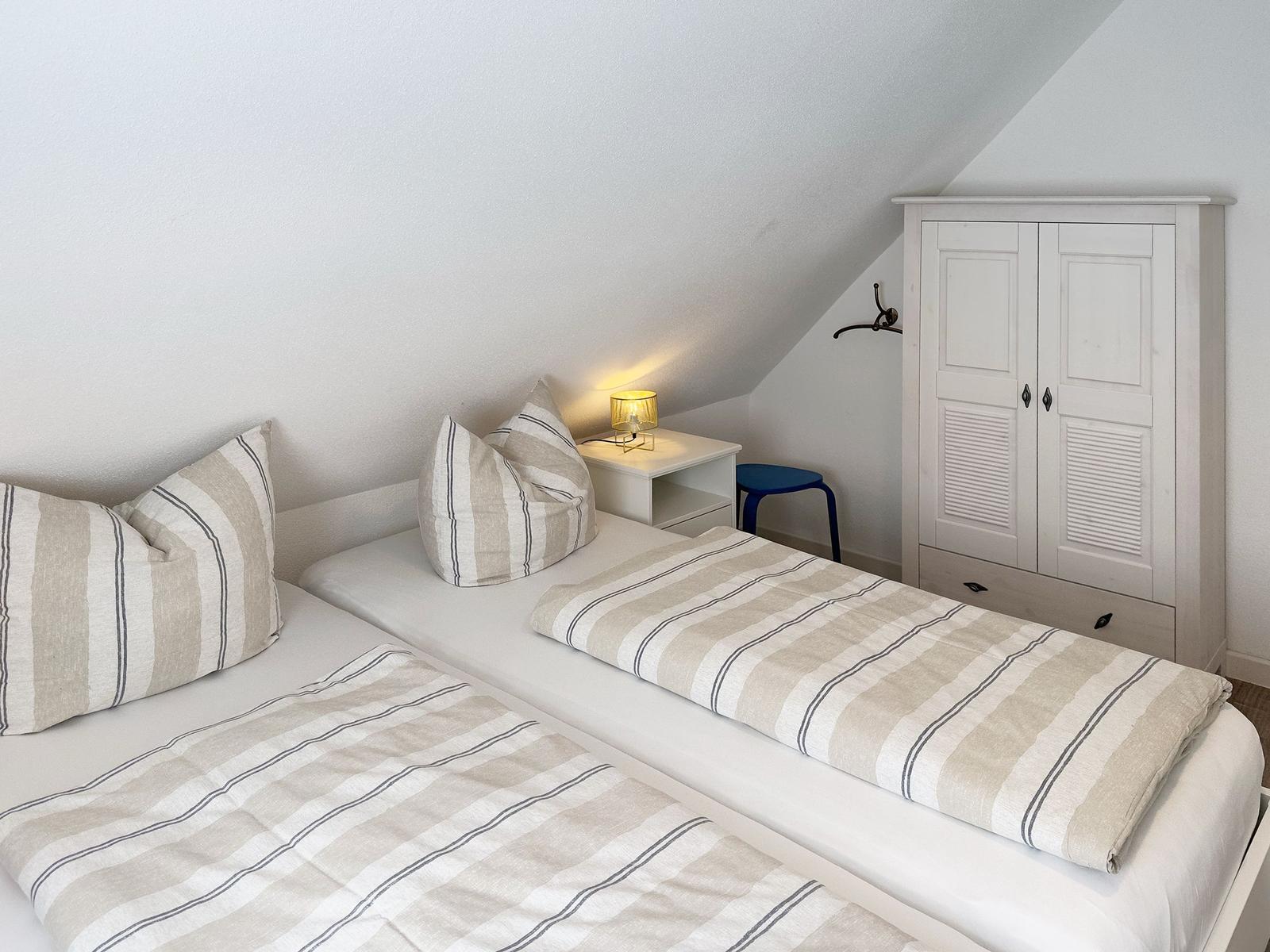 Two beds with striped bedding, nightstand with lamp, and white wardrobe.
