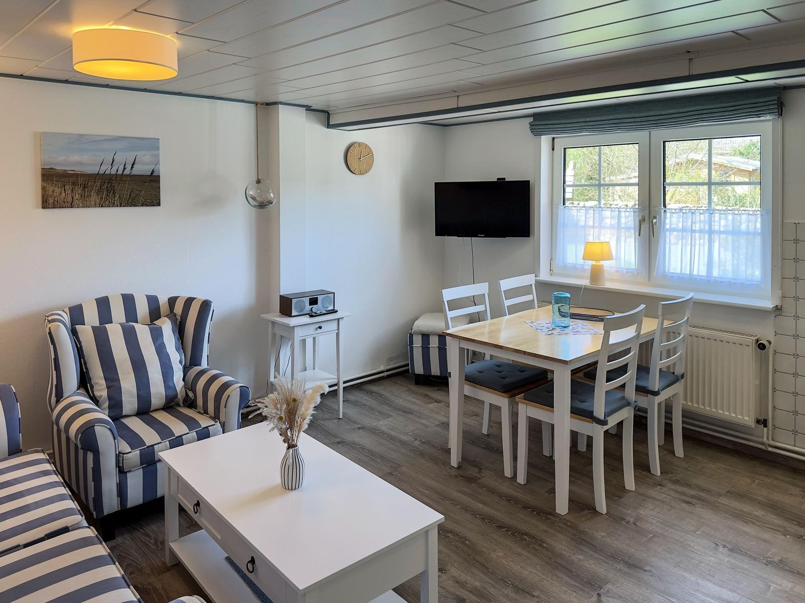 Wohn- und Essbereich mit Sofa, Tisch und Fernseher. Fenster mit Vorhängen.