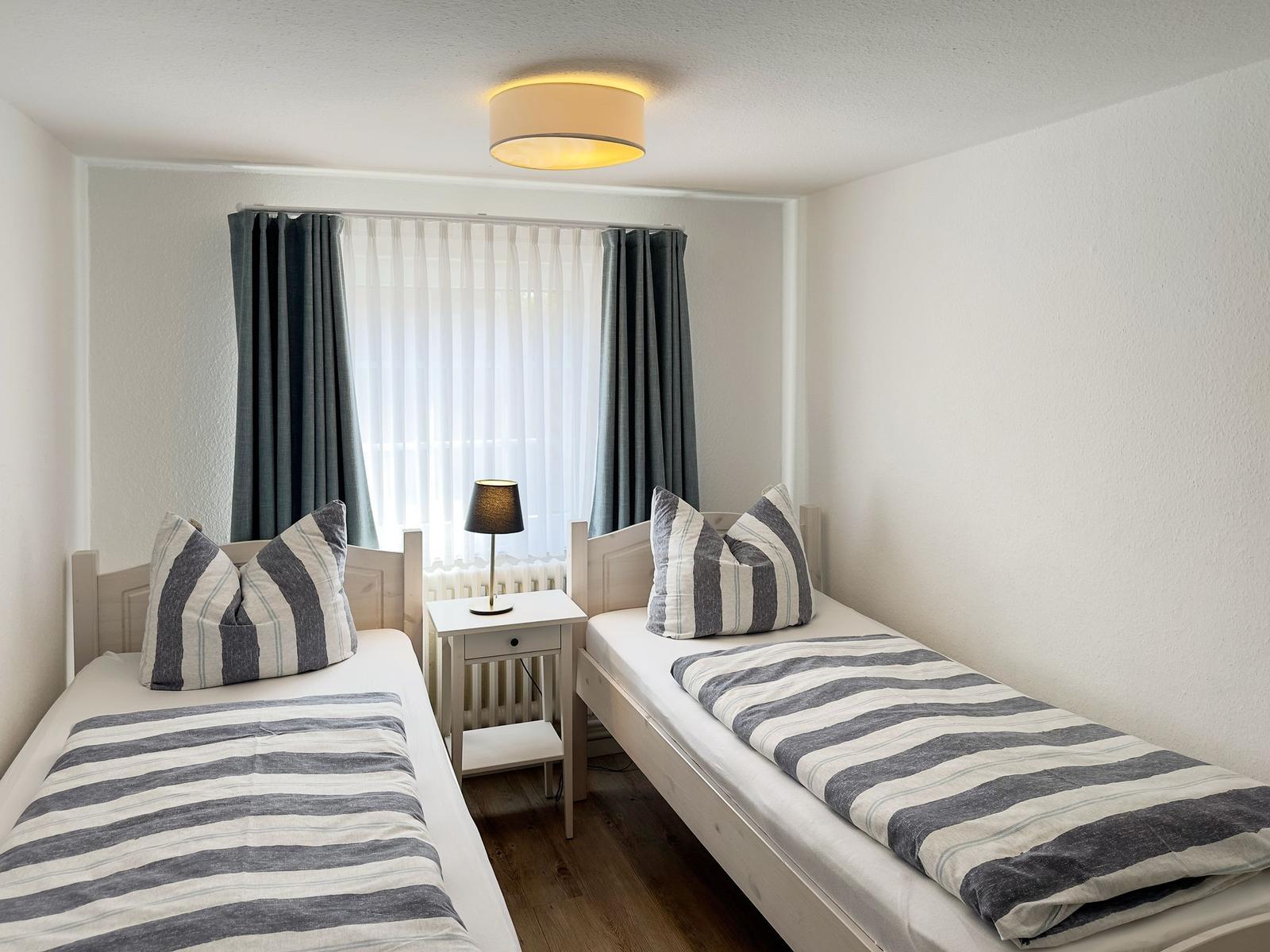 Zwei Einzelbetten mit gestreiften Bettwäsche und Kissen. Zwischen den Betten steht ein Nachttisch mit Lampe.