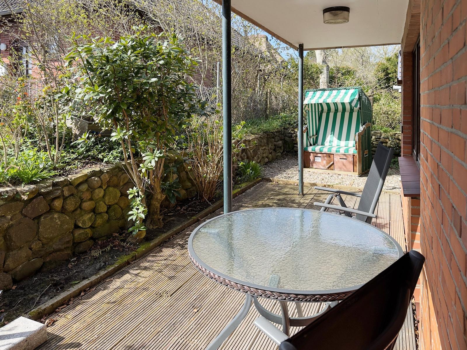 Terrace with table, chairs, and beach chair. Wooden deck, stone wall, and plants in the background.