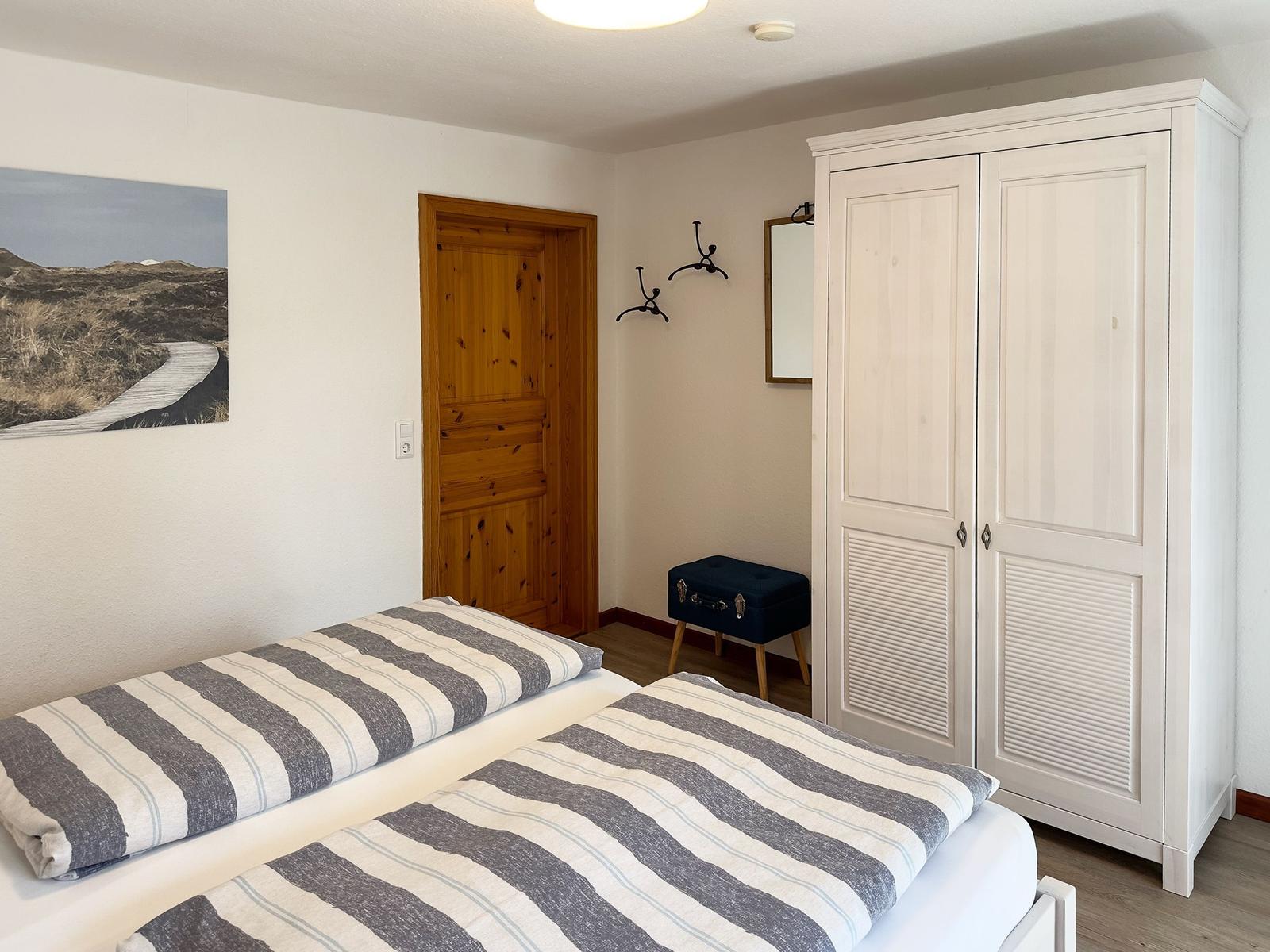 Two beds with striped bedding, white wardrobe, and wooden door.