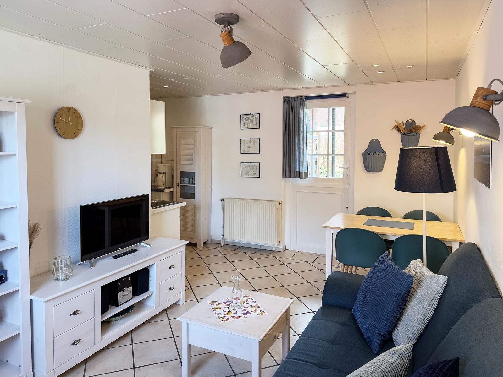 Living area with sofa, dining table, and TV. Kitchen in background. Window with curtains.