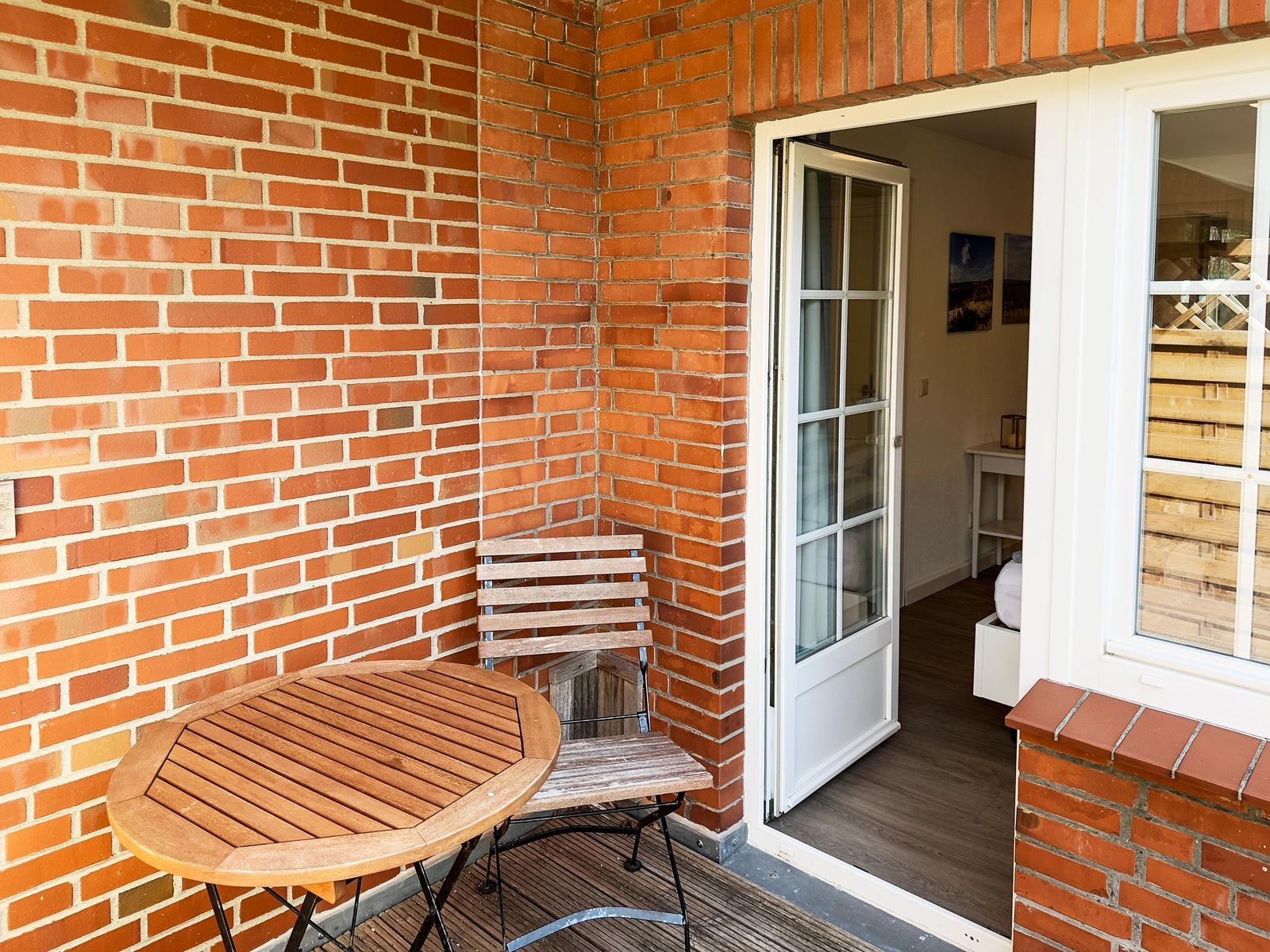 Terrasse mit Holztisch und Stuhl, rote Ziegelwand, Tür zum Innenraum.
