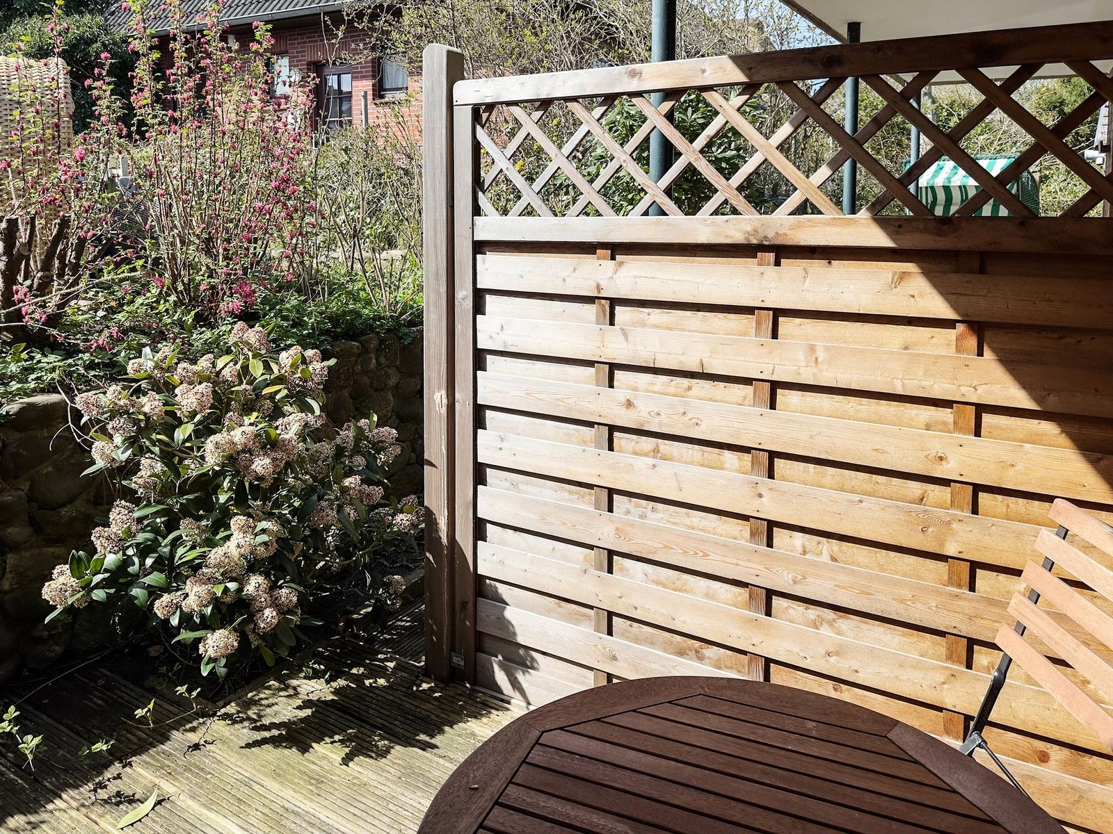 Holzterrasse mit Tisch, Sitzgelegenheit und Pflanzen. Holzgeländer mit Gitter oberhalb.