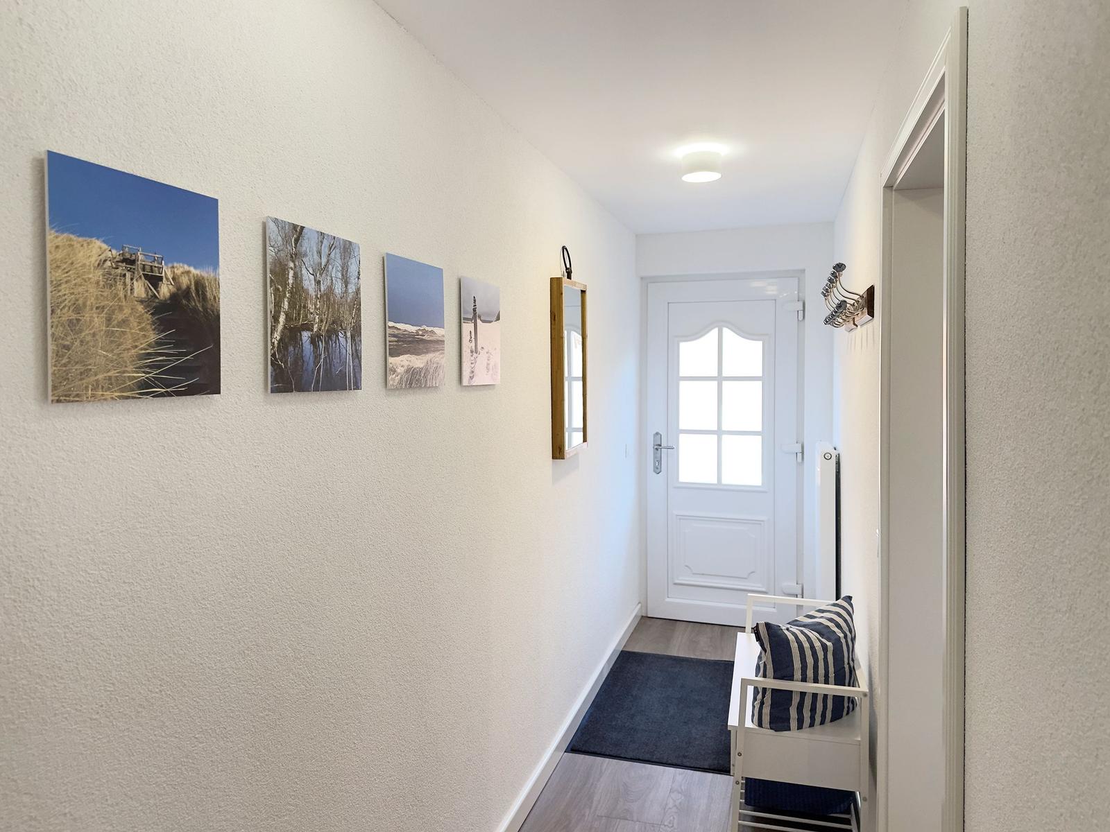 Flur mit Wandbildern, Spiegel, Tür und Sitzbank mit Kissen.