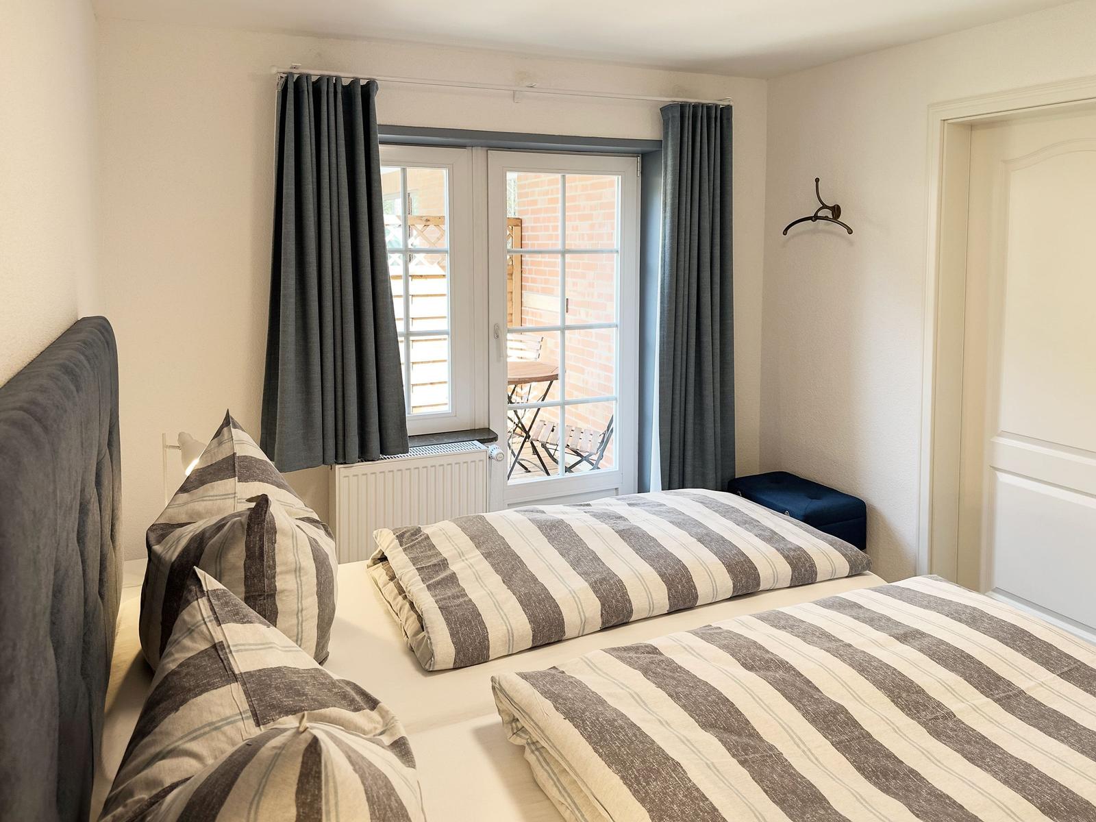 Schlafzimmer mit Doppelbett, Fenster und Vorhängen. Blick auf Balkon mit Tisch und Stühlen.