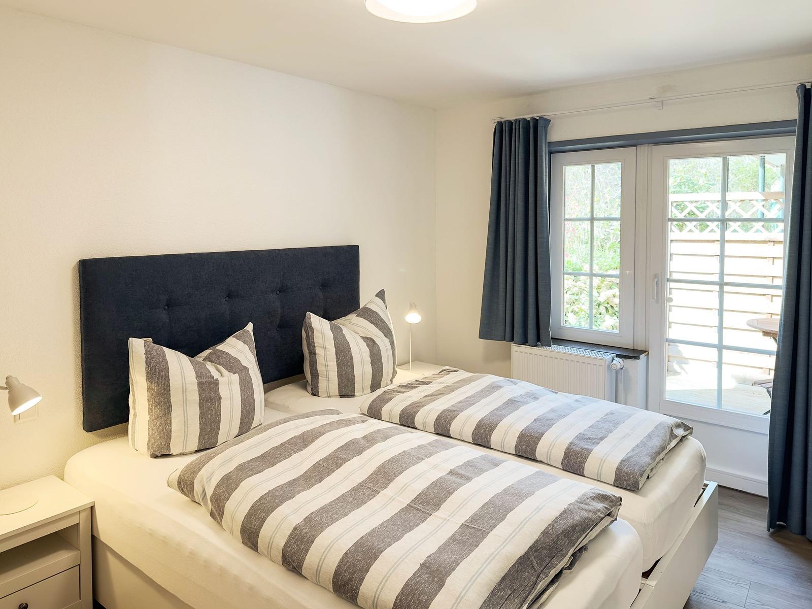 Schlafzimmer mit Doppelbett, Fenster und Vorhängen. Blick auf Balkon.