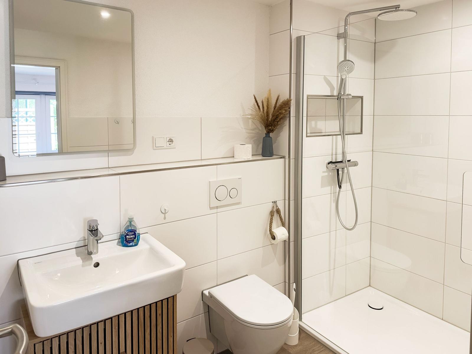 Badezimmer mit Dusche, Waschbecken und Toilette. Weiße Fliesen und Holzunterschrank.