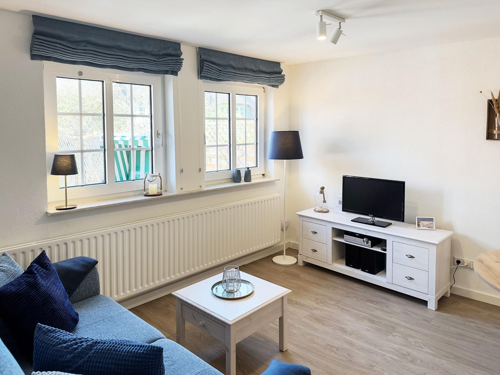 Wohnzimmer mit Sofa, Fernseher und zwei Fenstern mit blauen Vorhängen.
