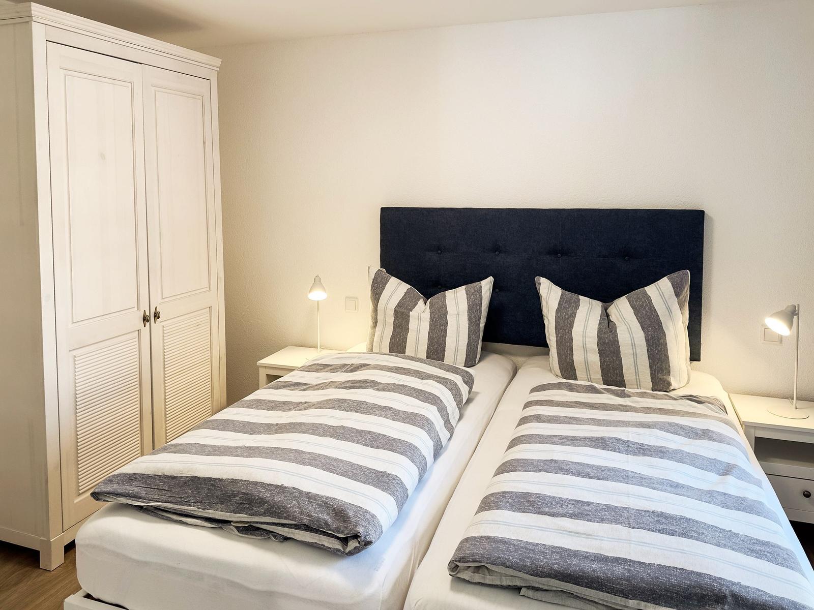 Zwei Betten mit Streifenbetten und dunklem Kopfteil, weiße Nachttische und Schrank.