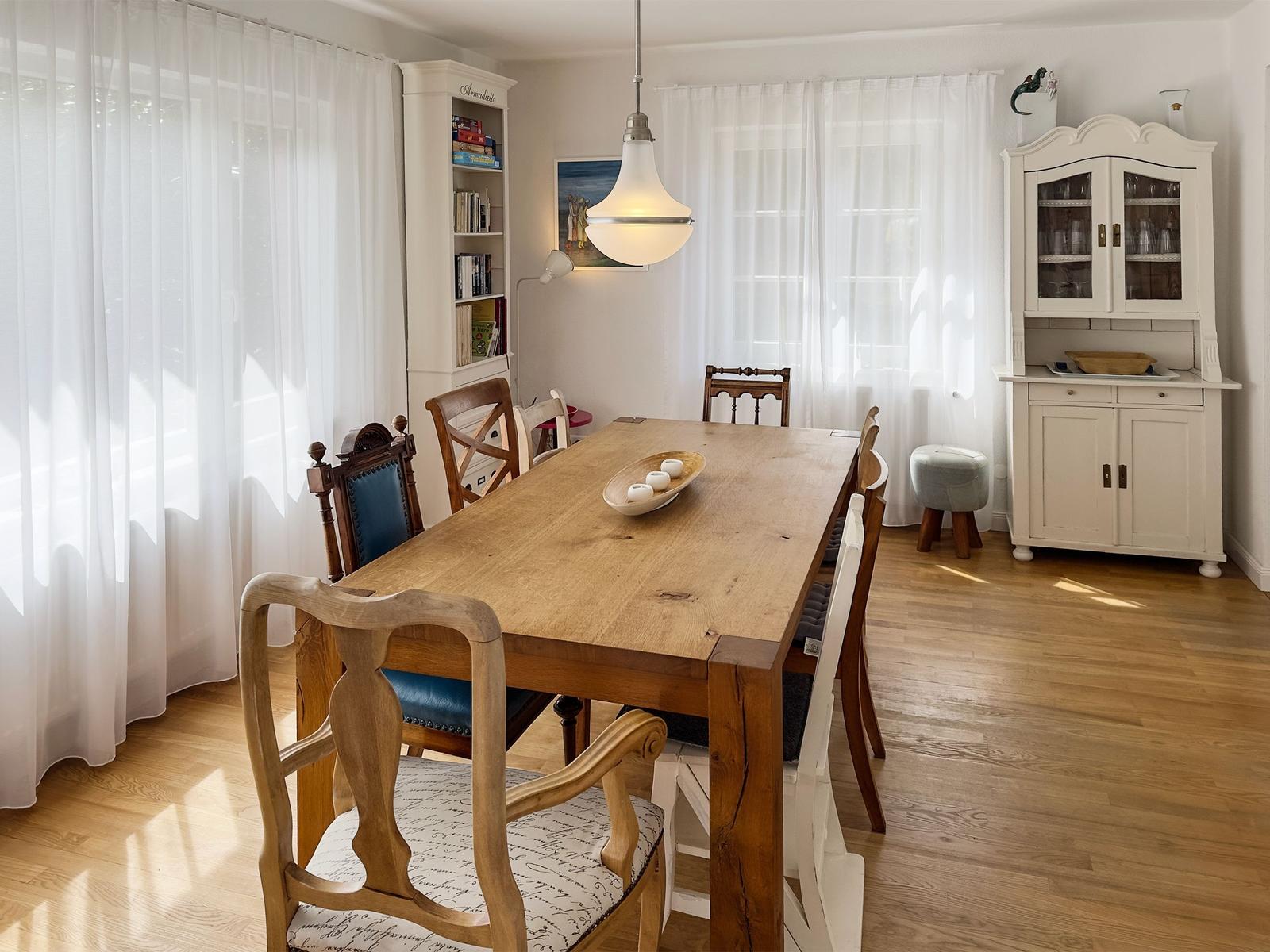Esszimmer mit Holztisch, Stühlen und weißen Schränken. Fenster mit weißen Vorhängen.