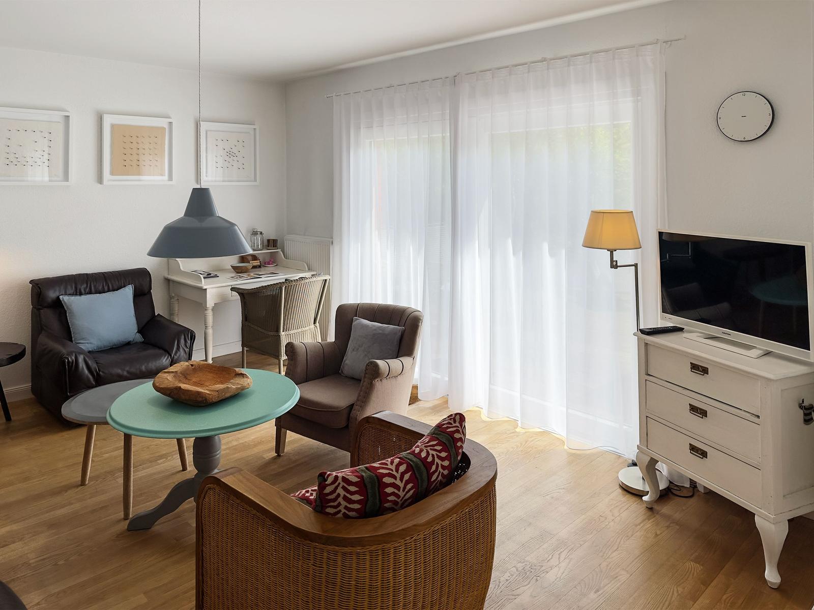 Wohnzimmer mit Sitzgelegenheiten, Schreibtisch, TV und großen Fenstern mit weißen Vorhängen.
