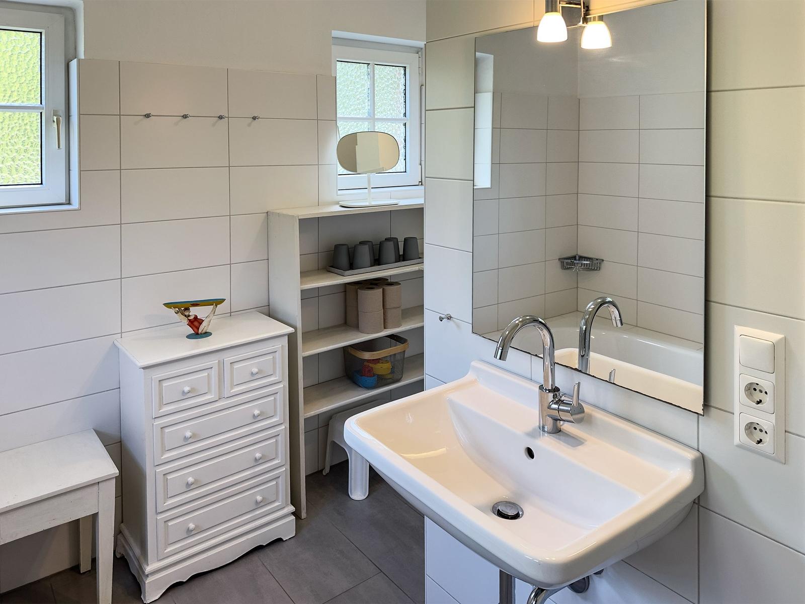 Badezimmer mit Waschbecken, Spiegel, Schrank und Regal. Fenster und Wandfliesen sichtbar.
