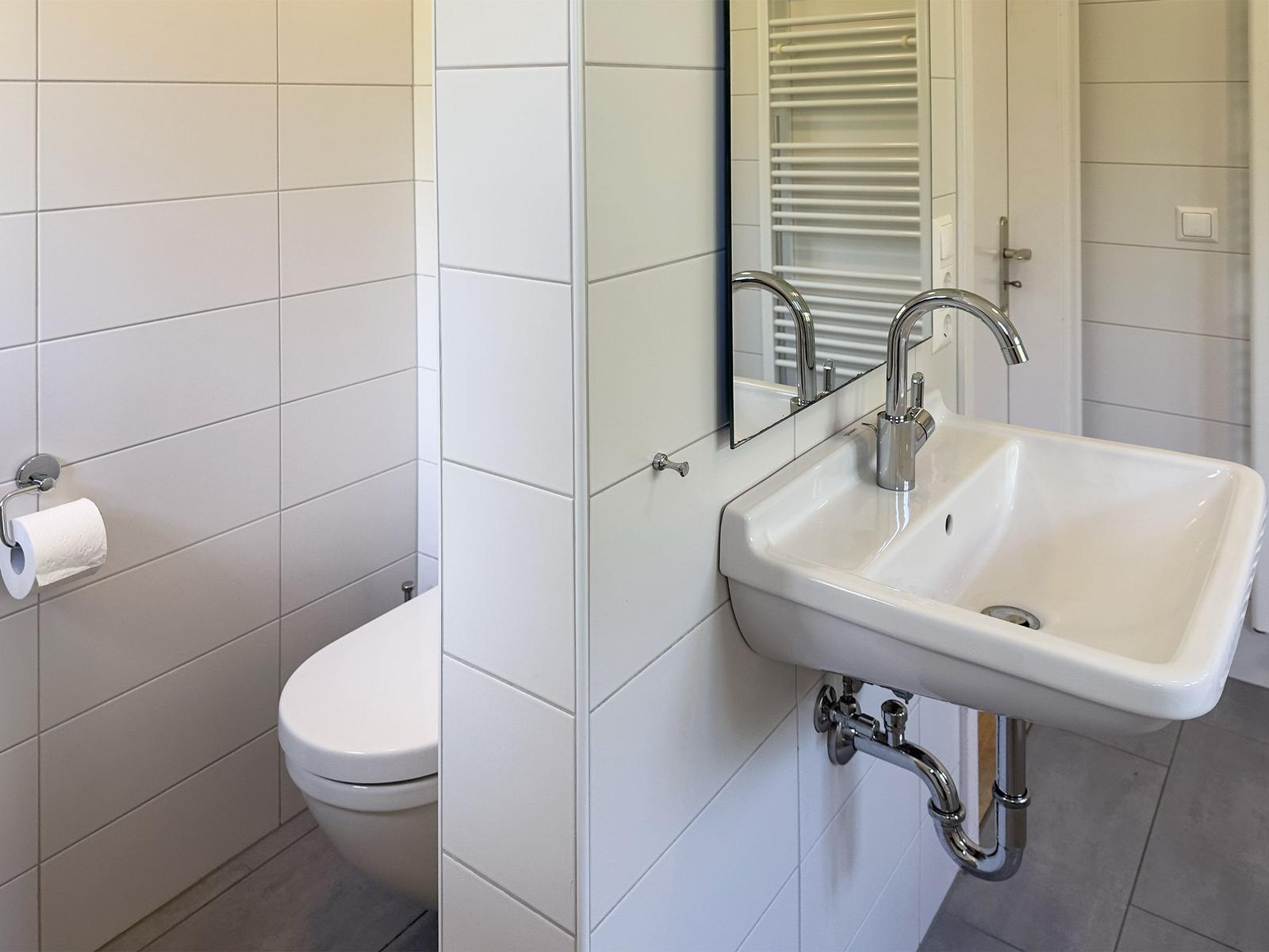 Badezimmer mit Toilette, Waschbecken und Spiegel. Fliesenwand und chromfarbene Armaturen.