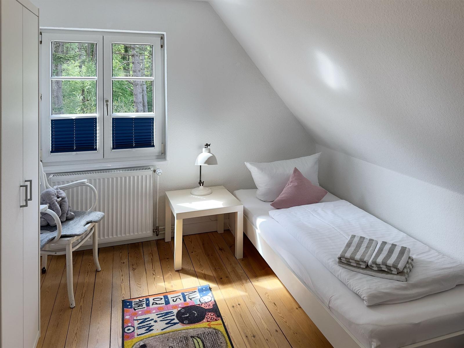Ein Schlafzimmer mit Dachschräge, Bett, Nachttisch, Stuhl und Fenster mit Blick auf Wald.
