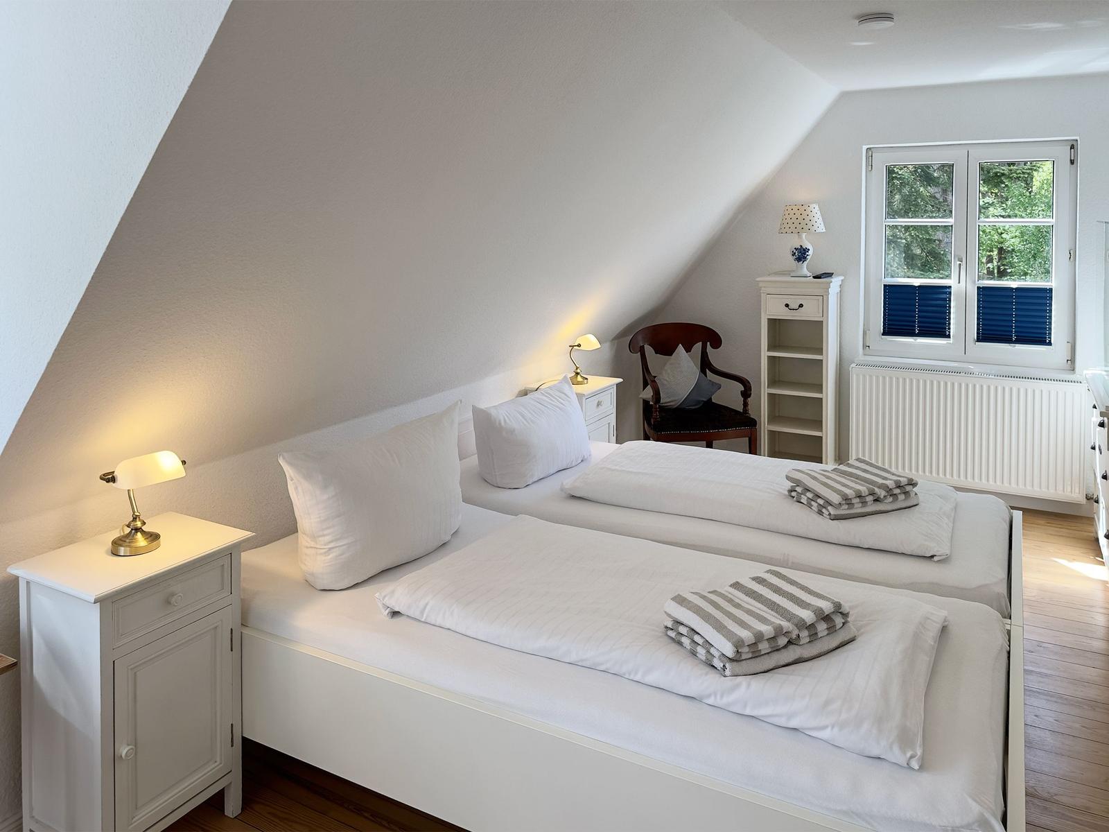Zwei Betten mit weißen Bettwäschen, Nachttische und ein Fenster mit Blick auf Bäume.
