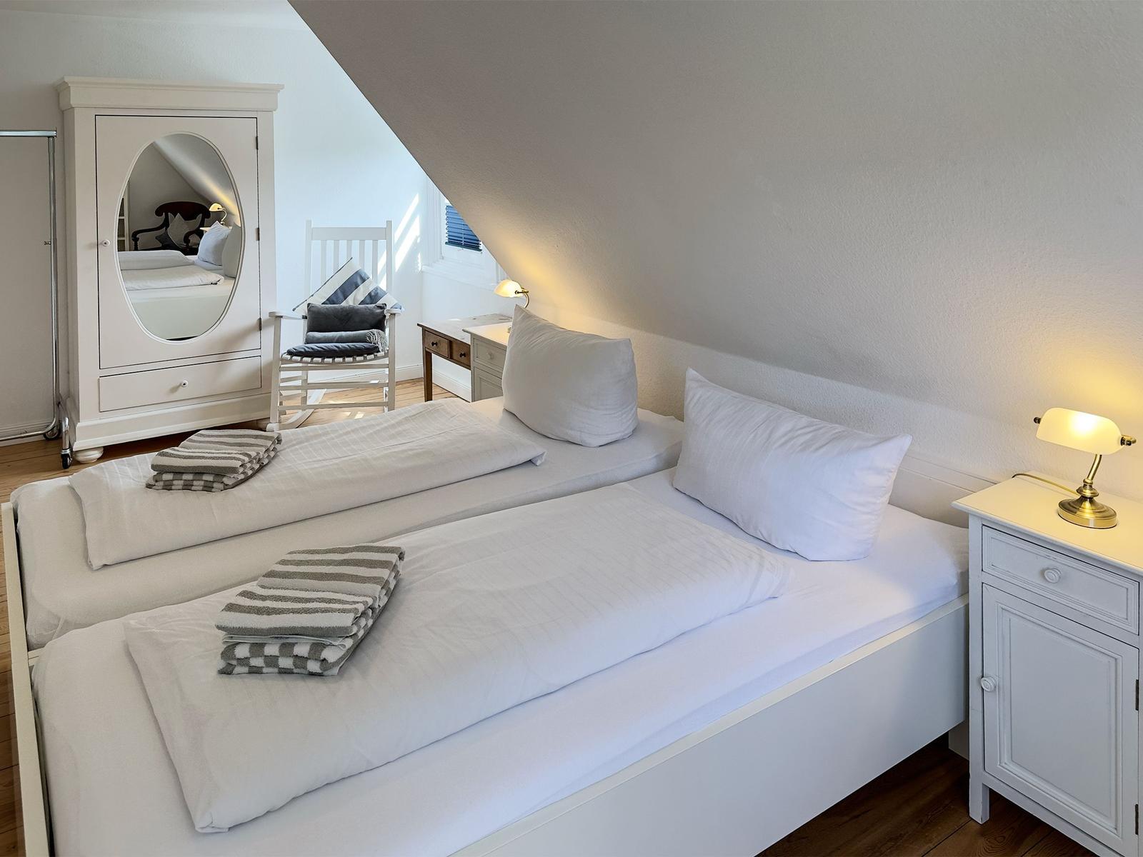 Zwei Betten mit weißen Laken und gestreiften Handtüchern. Weißer Schrank mit Spiegel und Nachttisch mit Lampe.