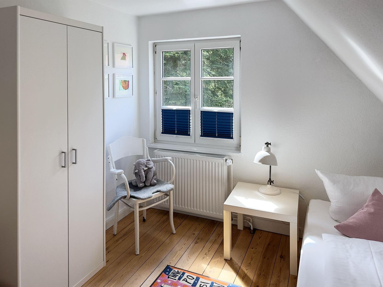 Zimmer mit Dachschrägen, Fenster, Schrank, Stuhl, Tisch und Bett.