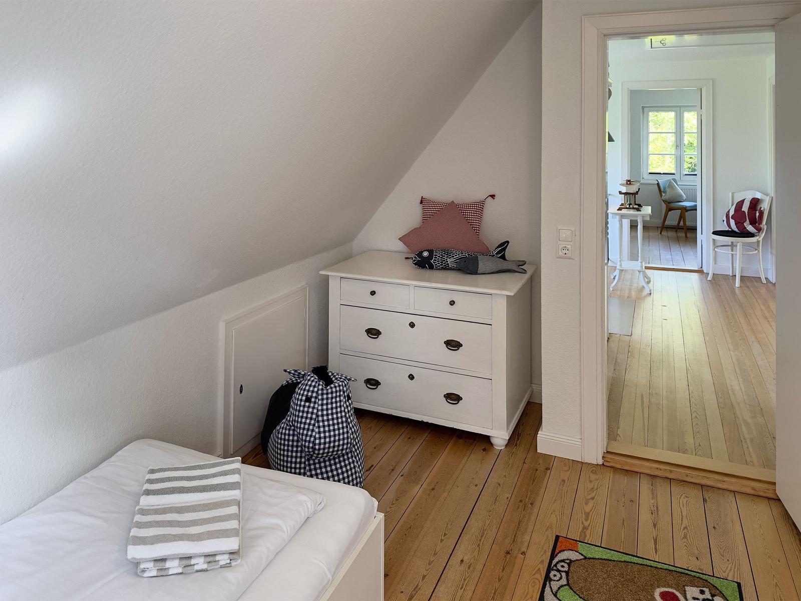 Schlafzimmer mit Dachschrägen, weißer Kommode und Blick in Wohnbereich.