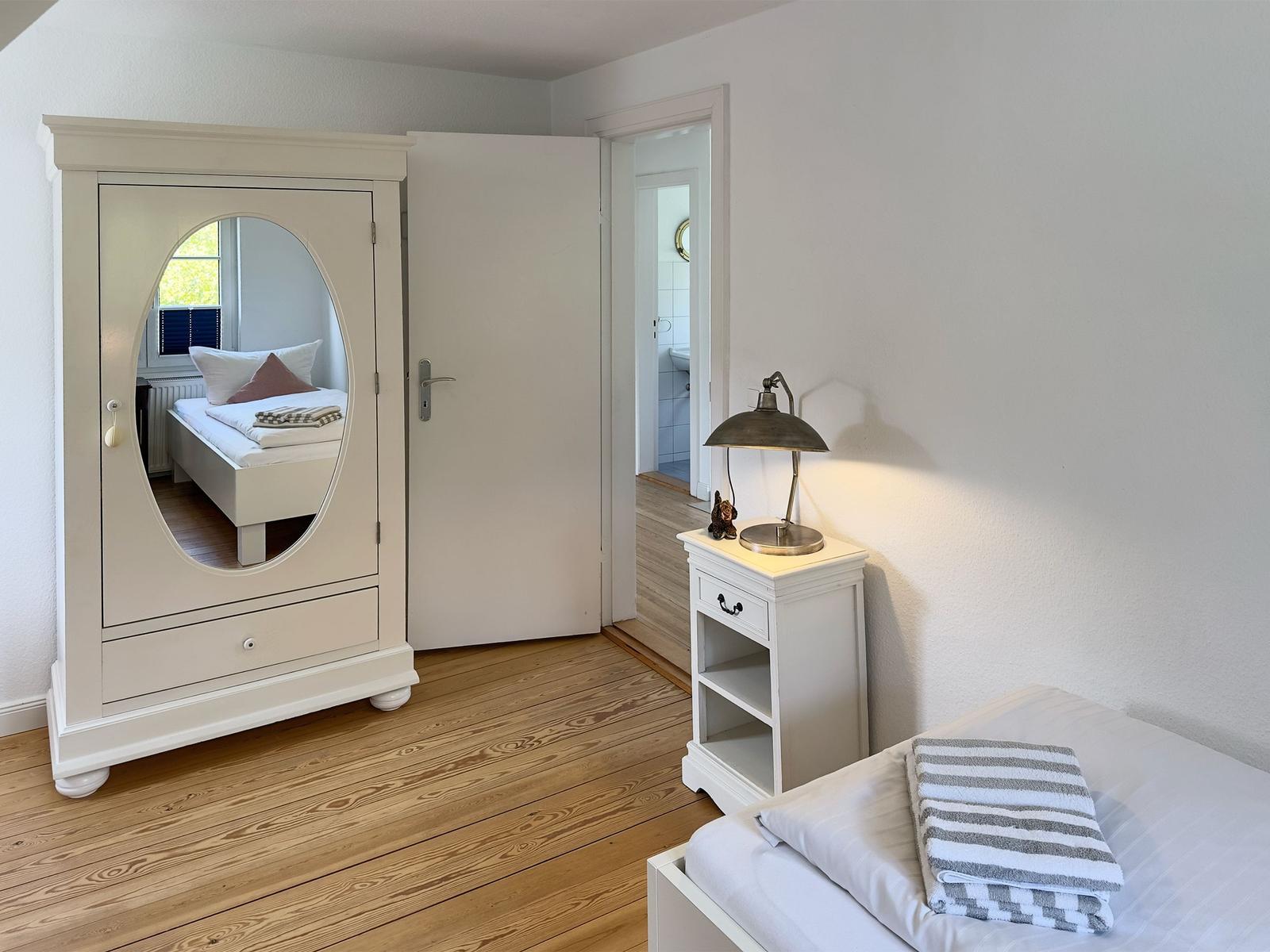 Schlafzimmer mit weißem Schrank, Bett und Nachttisch. Durchgang zur Badewanne.