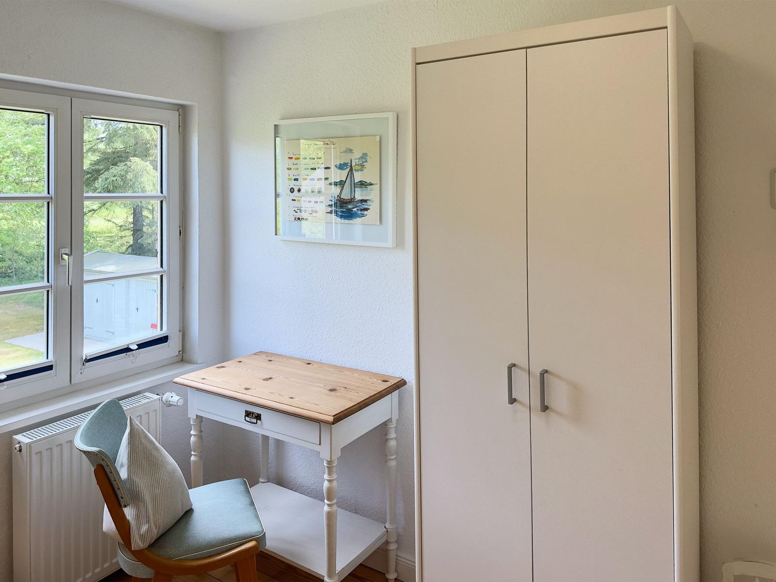 Schreibtisch mit Stuhl und Kleiderschrank neben Fenster mit Blick auf Garten.