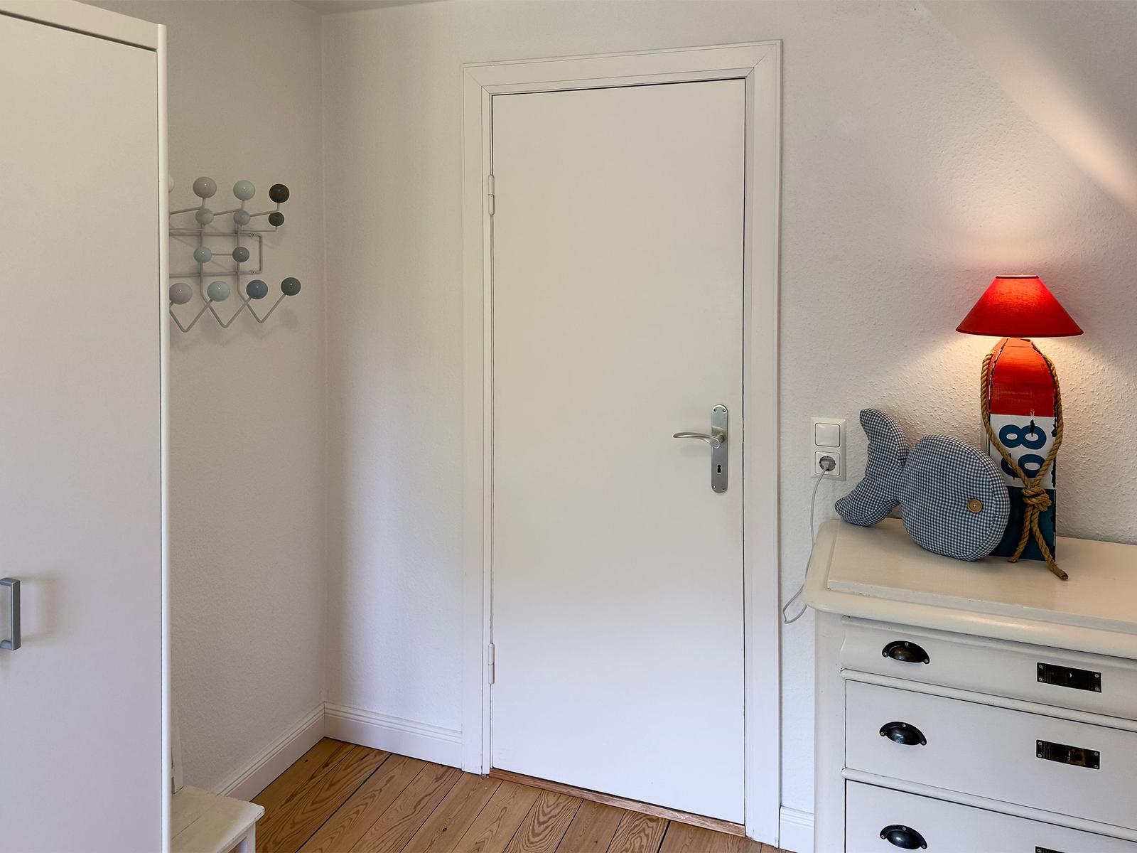 Ein Raum mit weißer Tür, Schrank und Nachttisch mit Lampe und Fischfigur.