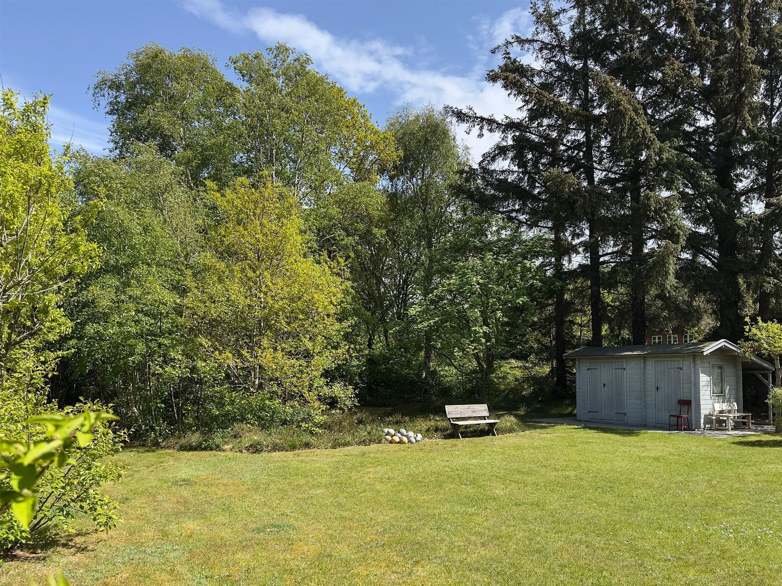 Grüner Garten mit Bank, Schuppen und Bäumen unter blauem Himmel.