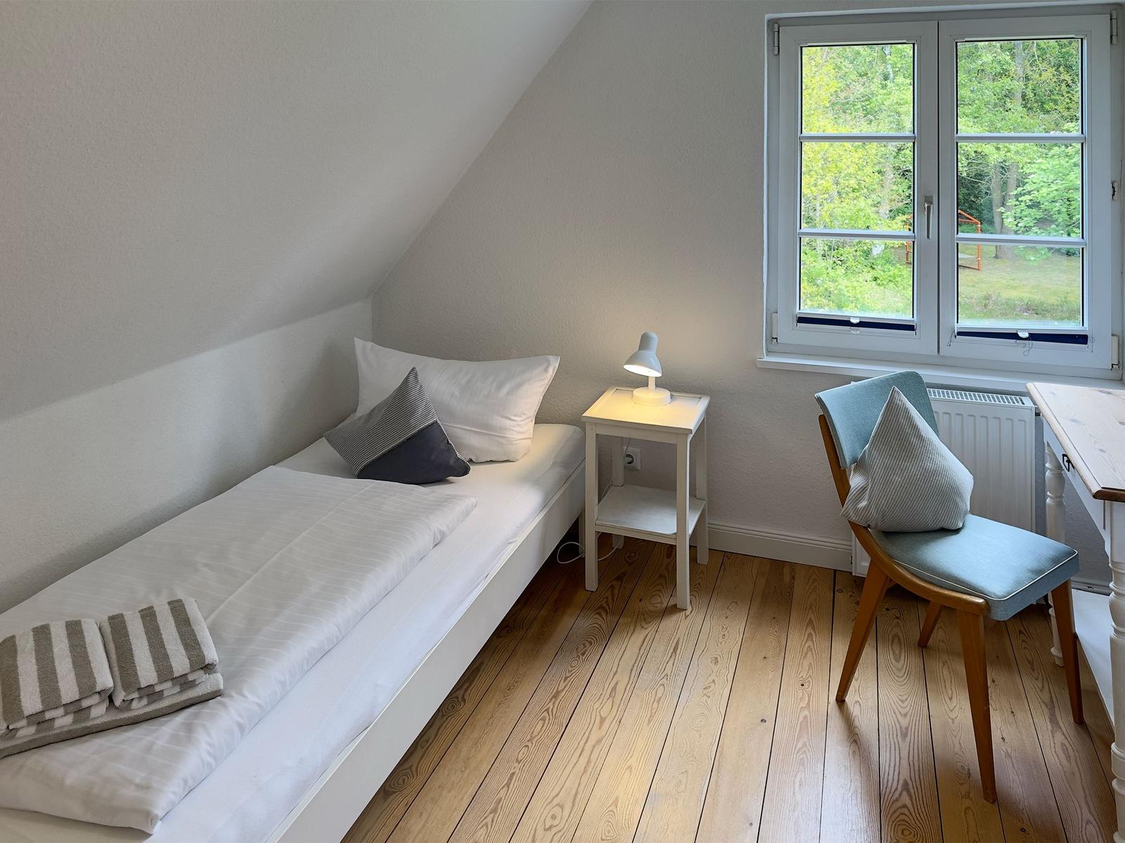 Ein Schlafzimmer mit Dachschrägen, Bett, Stuhl und Fenster mit Blick auf Grün.