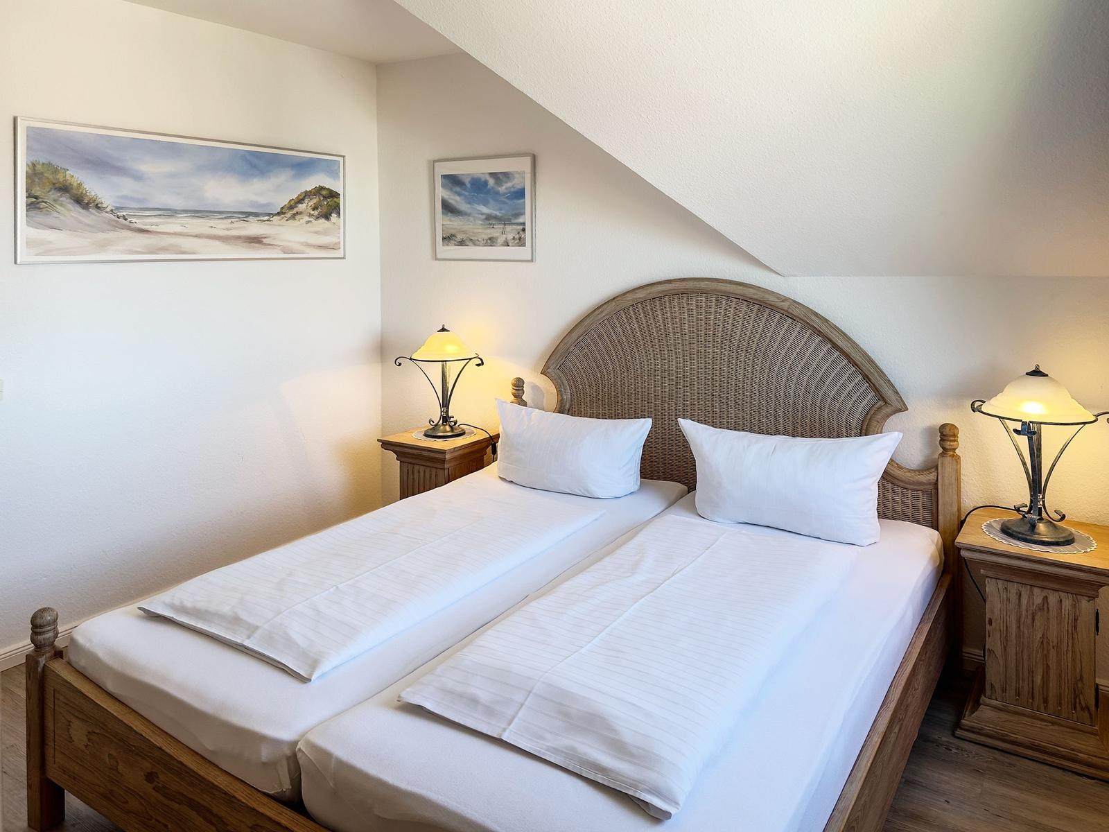 Zimmer mit Doppelbett, zwei Nachttischen und Lampen. Wandbilder zeigen Strandlandschaften.
