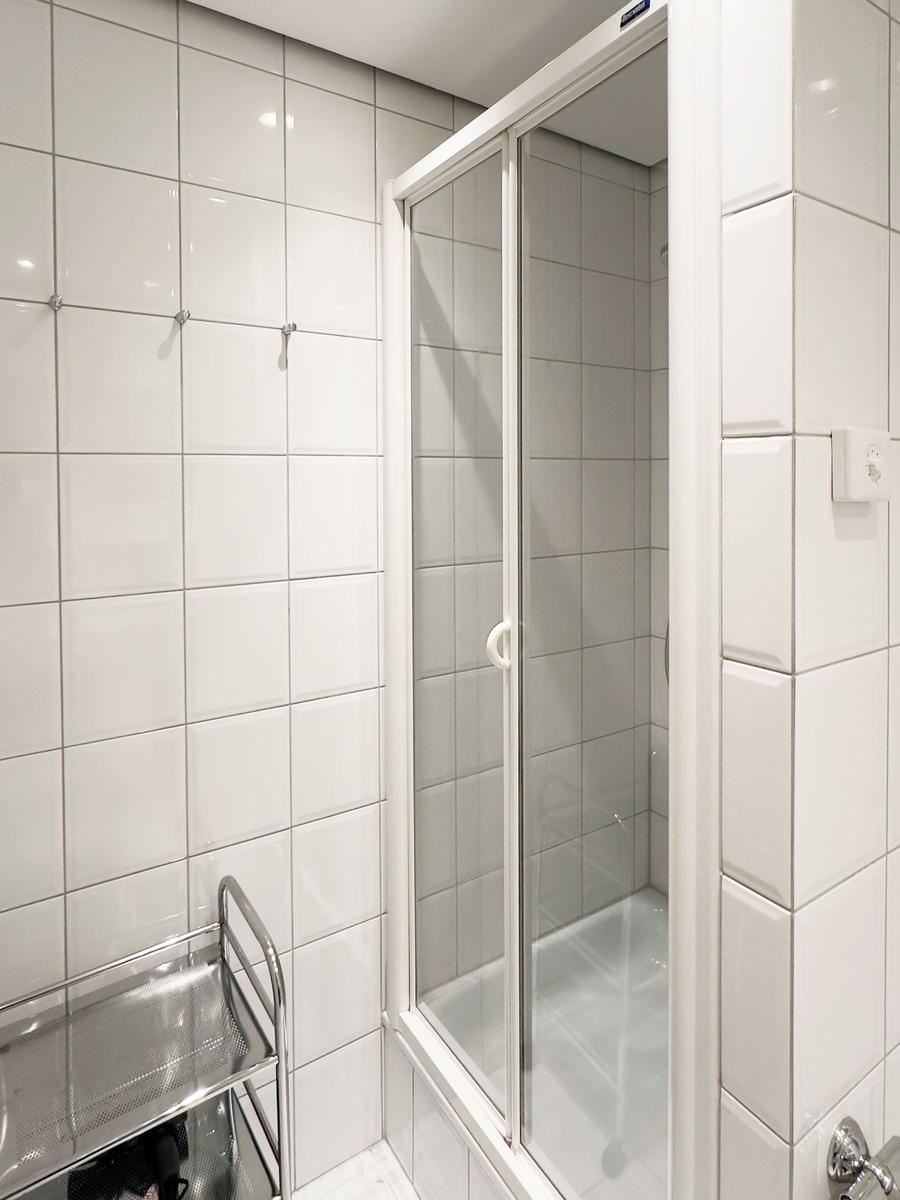 Dusche mit Glaswand und weißen Fliesen. Links steht ein Metallwagen.