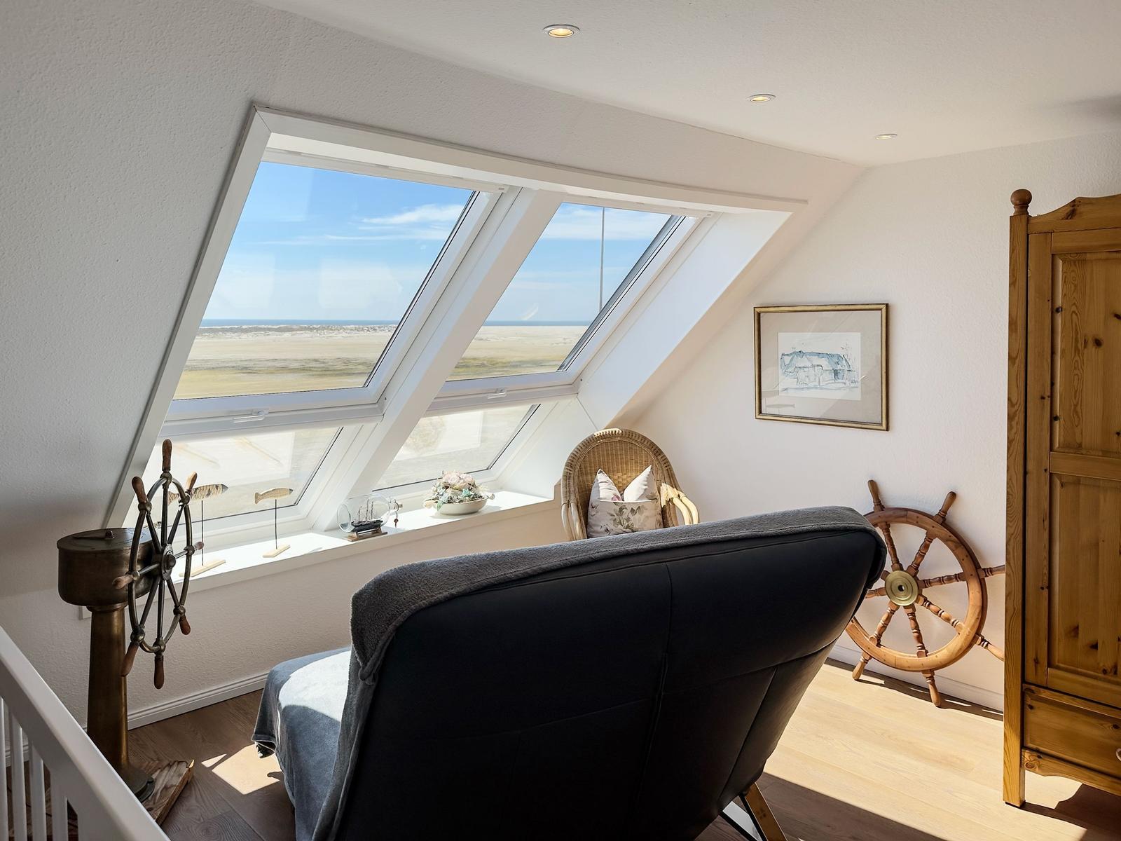 Dachgeschosszimmer mit großem Dachfenster, Blick auf Strand und Meer, Holzmöbel und Segelschiff-Deko.