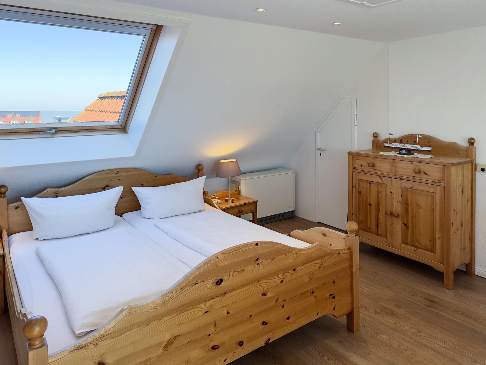 Schlafzimmer mit Holzbett, Schrank und Dachfenster mit Blick auf Dächer und Meer.